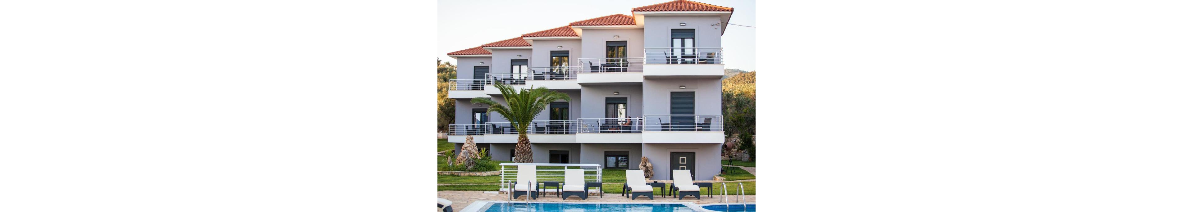 A contemporary three-story apartment building features multiple balconies and a terracotta tile roof. The setting includes a well-maintained garden with palm trees and a swimming pool in the foreground, surrounded by lounge chairs. The scene is brightly lit, evoking a relaxed and inviting vacation atmosphere, with no visible people or text.