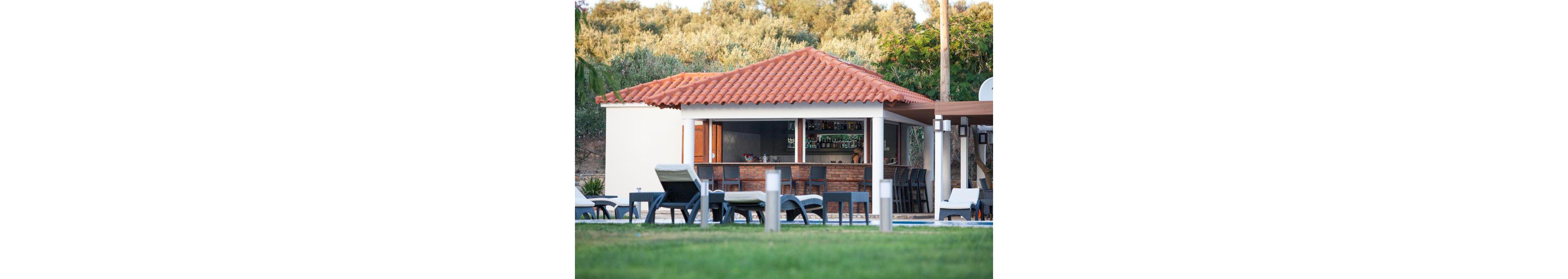 A small outdoor bar with a red tiled roof is situated beside a grassy area, featuring several lounge chairs and tables. The bar is open on one side, displaying shelves with bottles and glassware, and is surrounded by tall trees and greenery. The setting suggests a relaxed, vacation-like atmosphere, ideal for leisure and socializing. No visible text or numbers are present in the image.