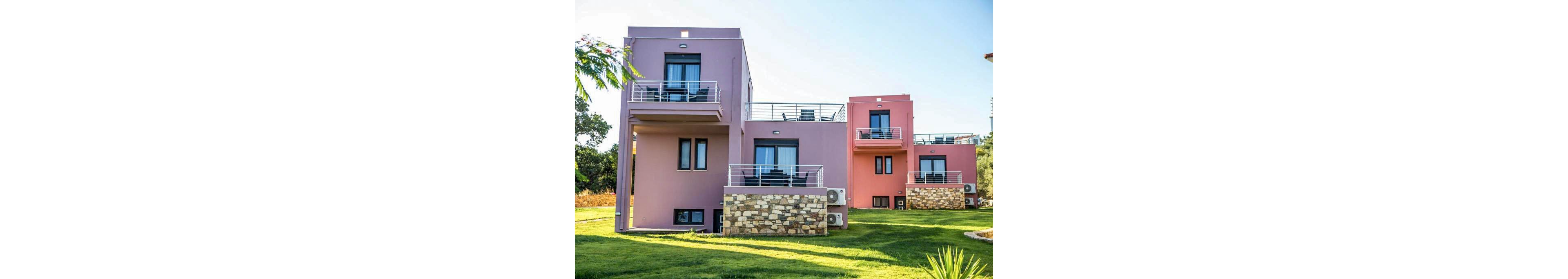 Three contemporary villas painted in pastel purple and pink tones stand side by side on a well-kept grassy lawn. Each building features private balconies with metal railings, large windows, and stonework on the lower facade. The setting is bright and sunny, suggesting a peaceful residential or vacation area. Air conditioning units are visible on the exterior walls, and the overall mood is calm and inviting.