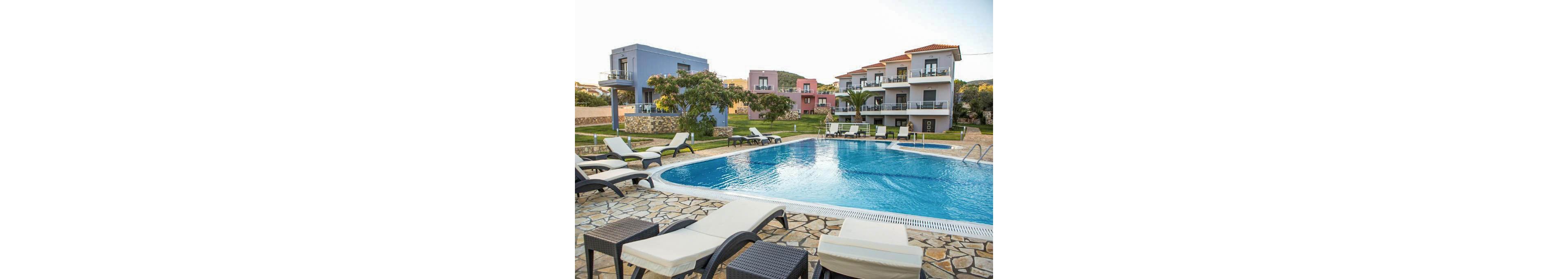 A serene outdoor swimming pool is surrounded by several modern apartment buildings with balconies. Multiple sun loungers and small tables are arranged around the pool on a stone-tiled deck, creating a relaxing vacation atmosphere. The setting is bright and inviting, with clear blue water and well-maintained green lawns. No visible people or text are present in the image.