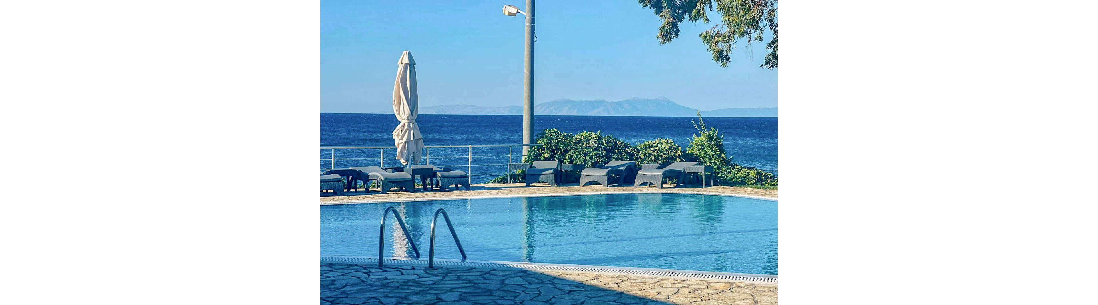 A tranquil swimming pool is set beside a stone deck, overlooking a calm blue ocean with distant mountains on the horizon. Several lounge chairs and a closed umbrella line the poolside, suggesting a relaxing atmosphere. The scene is bathed in natural daylight, with clear skies and lush greenery adding to the peaceful mood. No people are present, emphasizing the quiet and inviting setting.
