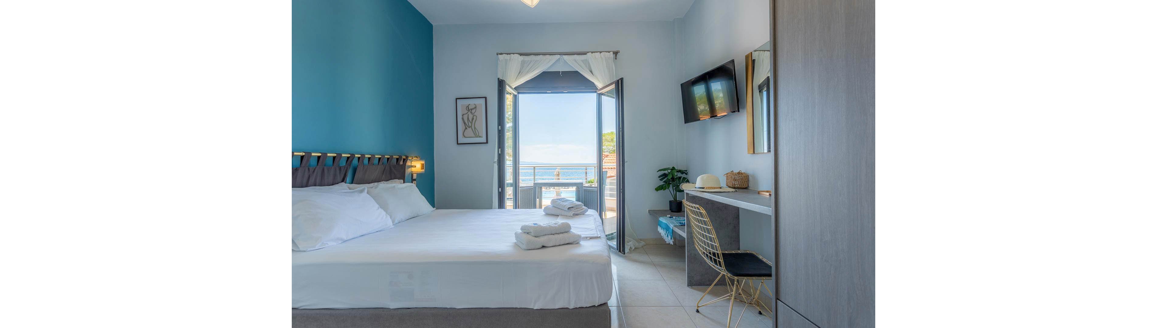 A contemporary hotel room features a large bed with crisp white linens and folded towels, facing a balcony with a scenic ocean view. The room includes a desk with a chair, wall-mounted television, and minimalist decor in cool blue and neutral tones. Natural light fills the space, creating a fresh and inviting atmosphere, with a hat and plant adding subtle personal touches.