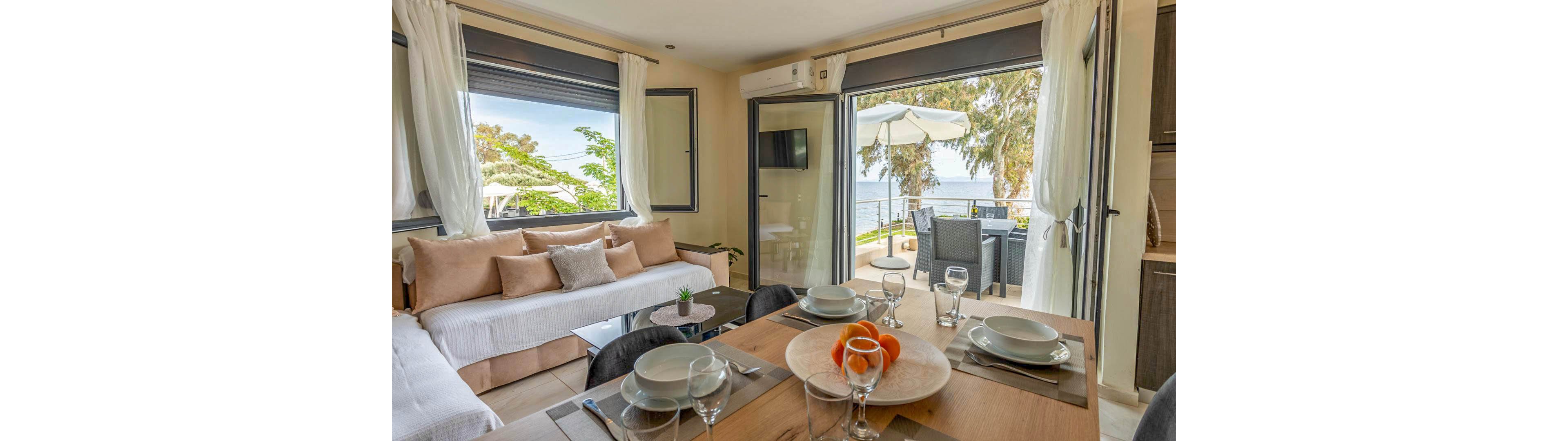A bright, modern apartment features a cozy living area with beige cushions and a dining table set for four. Large windows and glass doors open to a balcony with outdoor seating and a white umbrella, overlooking the sea and lush greenery. The space is decorated in neutral tones with natural light, creating a relaxed and inviting atmosphere. Tableware and a bowl of oranges add a touch of homeliness to the setting.