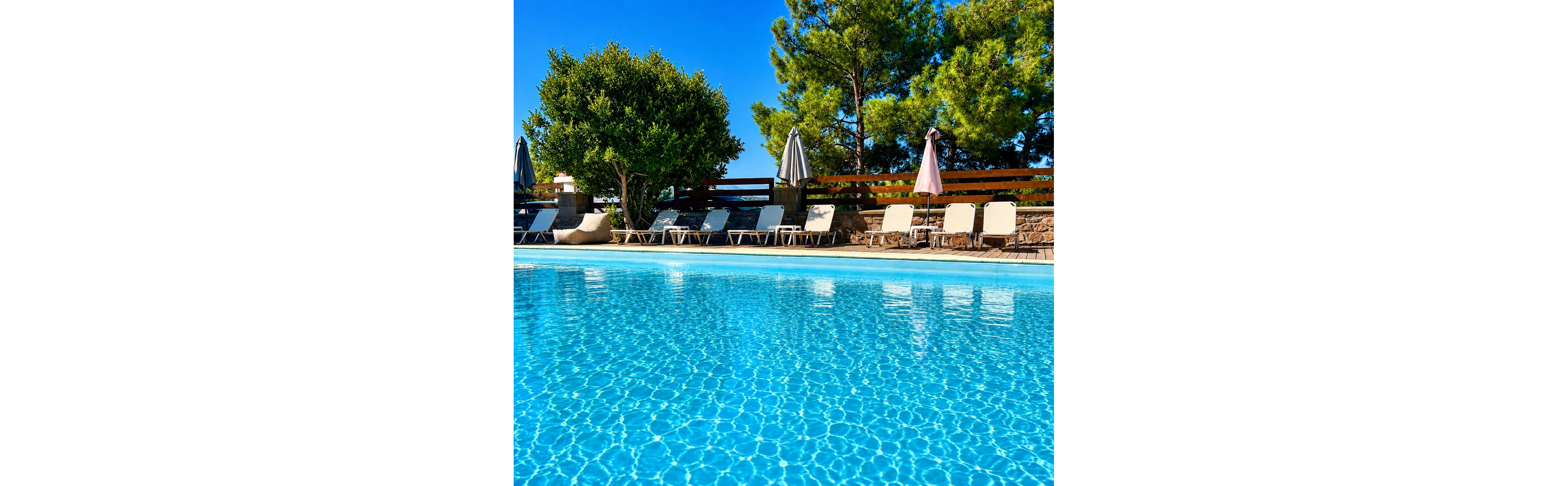 A clear blue swimming pool sparkles under bright sunlight at an outdoor resort. Several sun loungers line the poolside, accompanied by closed umbrellas and shaded seating. Tall green trees and a wooden fence form a natural backdrop, creating a relaxing vacation atmosphere. The scene suggests a tranquil, warm day ideal for leisure and sunbathing.