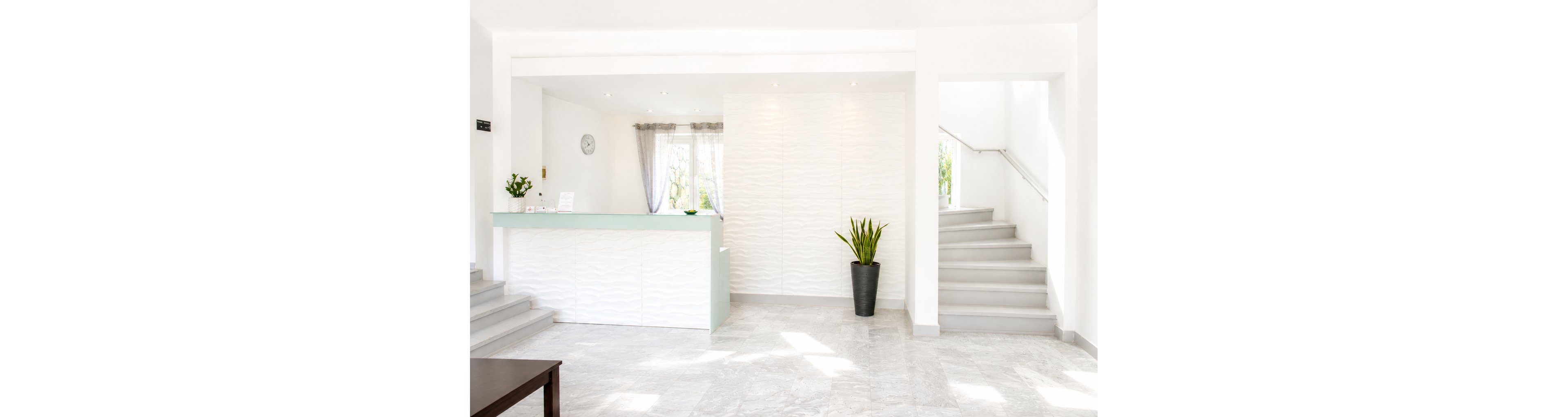 A modern reception desk stands in a bright, minimalist interior. The space features white textured walls, light stone flooring, and a frosted glass-topped counter. A potted plant adds a touch of greenery beside the desk, while a staircase on the right leads to an upper level. Natural light streams through sheer curtains, creating a clean and welcoming atmosphere.