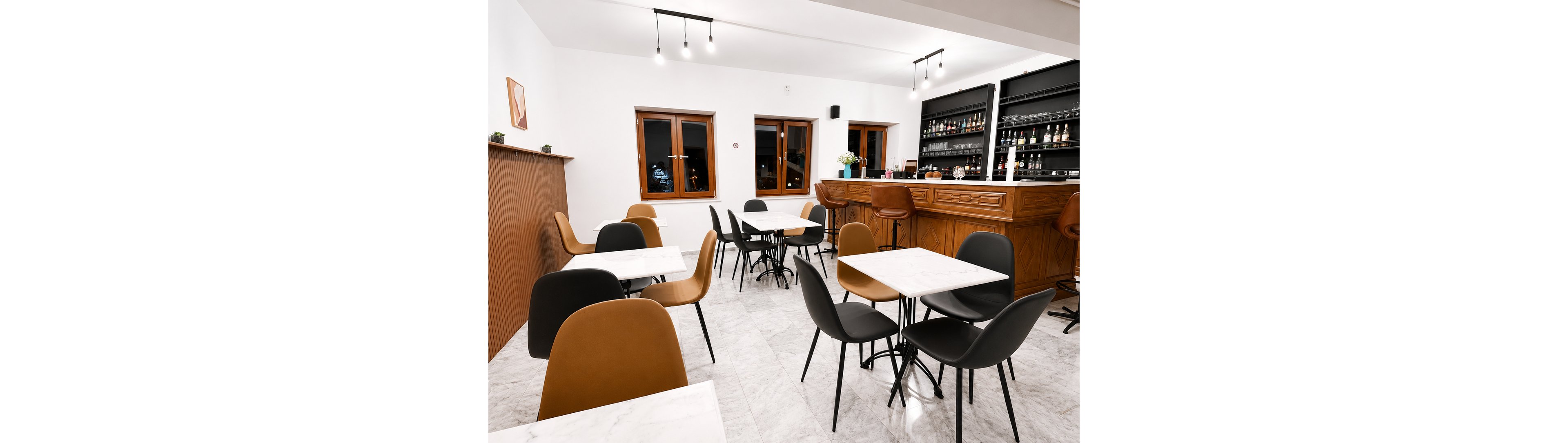 A contemporary café interior features marble-top tables and mixed black and tan chairs. The space is empty, showcasing a clean and minimalist dining area. In the background, a wooden bar counter with shelves of bottles and glassware adds warmth and character. White walls, tiled flooring, and simple ceiling lights create a bright, modern atmosphere.
