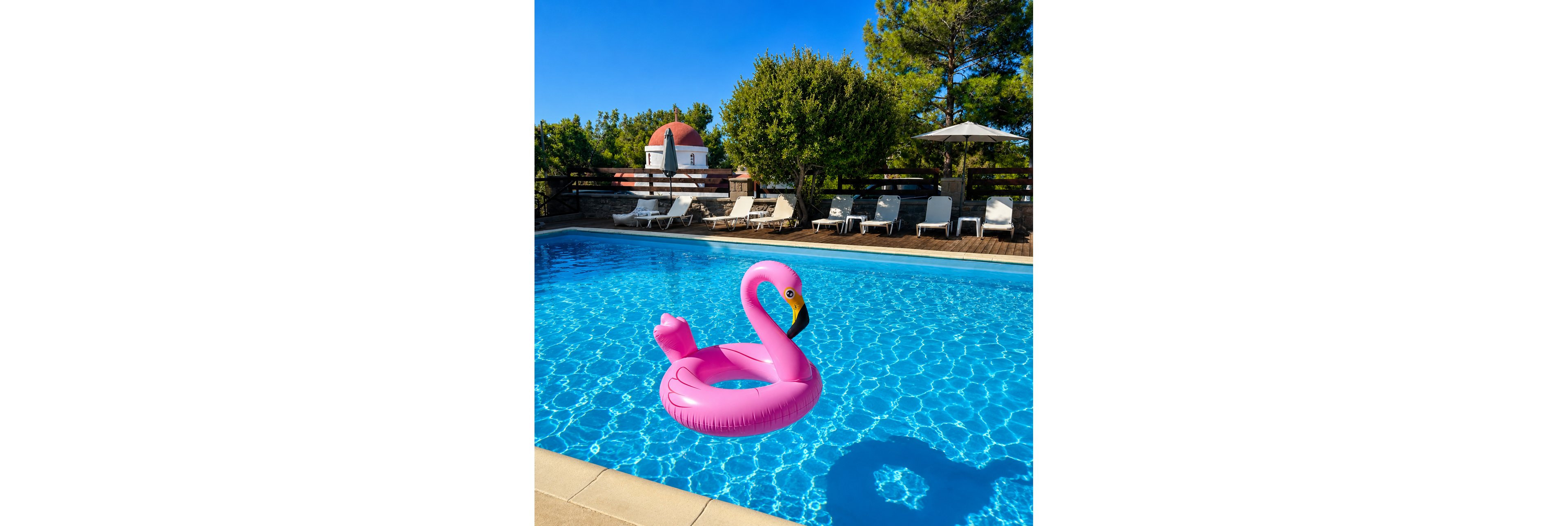 A bright pink flamingo inflatable float drifts in a clear blue swimming pool. The pool is surrounded by white lounge chairs and umbrellas on a wooden deck. In the background, trees and a small building with a red dome roof are visible under a clear blue sky. The scene suggests a relaxing summer vacation atmosphere at a resort or villa.
