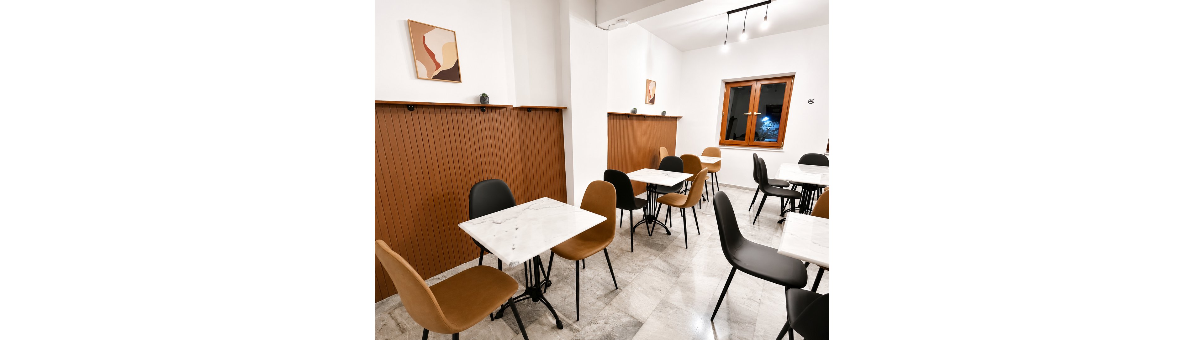 A modern cafe interior features marble-top tables paired with black and tan chairs. The space has clean white walls, warm wooden paneling, and simple abstract artwork. Large windows bring in natural light, while ceiling lights provide additional illumination. The overall design feels minimalist, tidy, and inviting for guests.