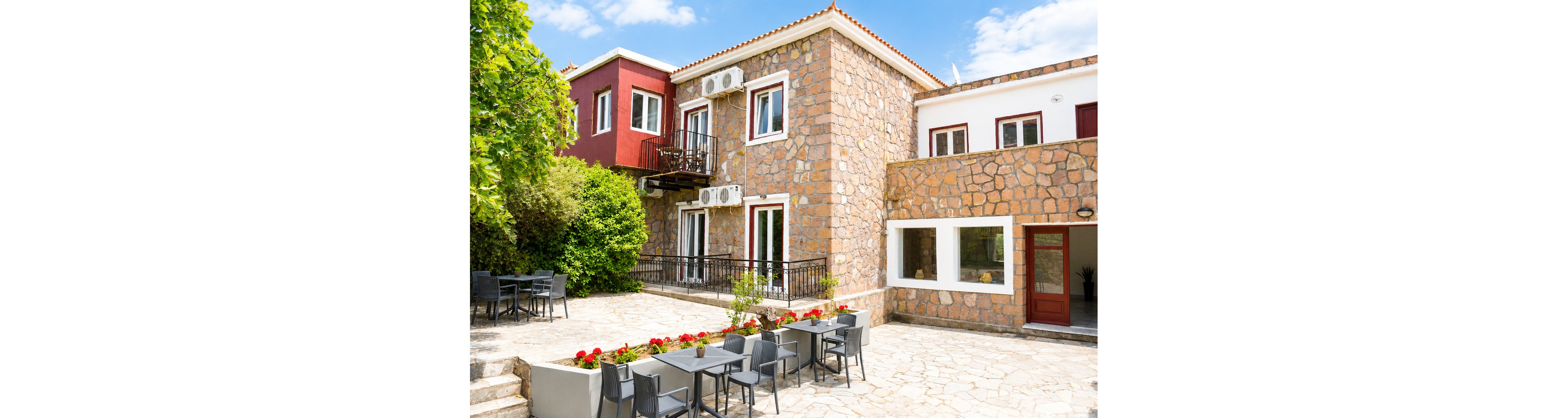 A two-story stone guesthouse features multiple windows, balconies, and exterior air conditioning units. The building opens onto a spacious stone-paved courtyard with several tables and chairs arranged for outdoor dining or relaxation. Bright red flowers in planters add color to the neutral stone tones. Lush greenery borders one side of the courtyard, and the sky above is clear with scattered clouds, suggesting a pleasant, sunny day.