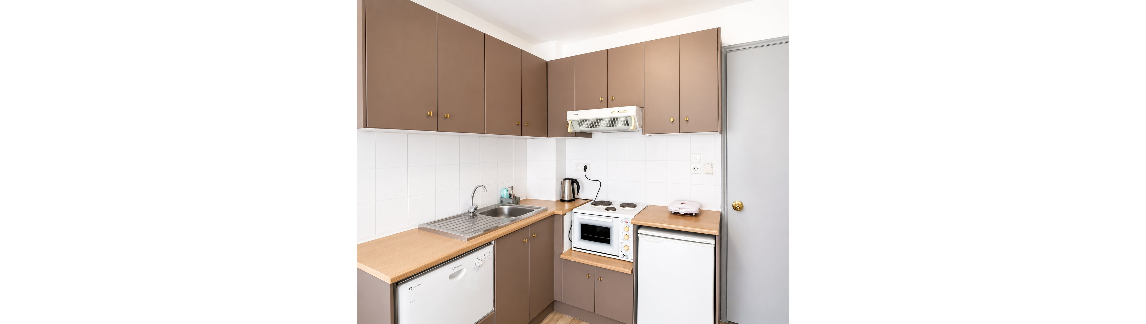 A small, tidy kitchen features brown cabinets and light wood countertops. The space includes a stainless steel sink, electric stove, countertop oven, kettle, and a compact refrigerator. White tiled walls and a neutral door with a brass knob add to the clean, minimalist look. The overall scene suggests a functional apartment or studio kitchen designed for everyday cooking.