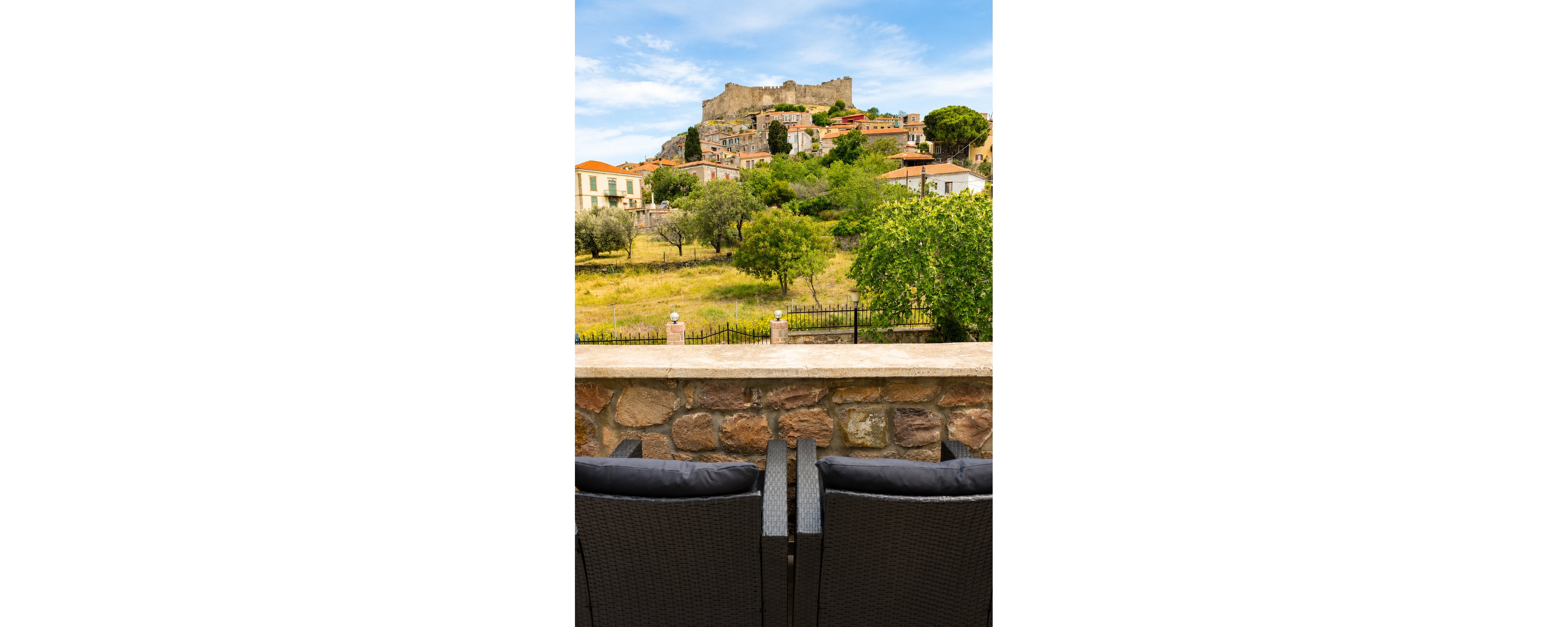 A pair of dark wicker chairs on a stone terrace overlook a lush green field and a charming hillside village. Colorful houses with tiled roofs climb the slope toward an ancient stone castle crowning the hill. Trees and shrubs dot the landscape, adding vibrant greenery to the scene. A bright blue sky with scattered clouds enhances the peaceful, picturesque atmosphere.