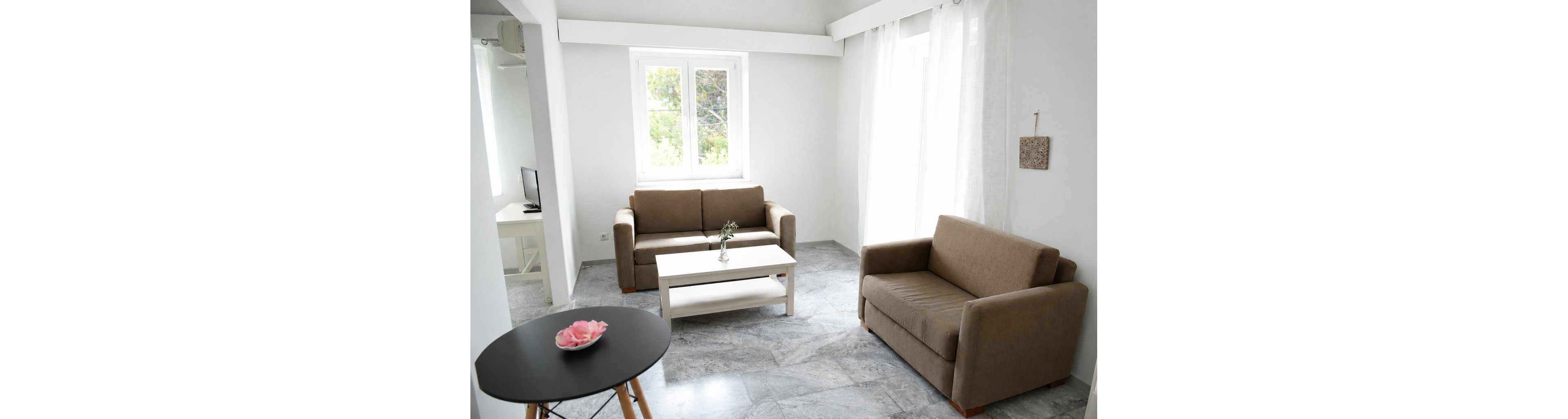 A bright minimalist living room features two brown sofas facing a white coffee table. Large windows and sheer curtains allow natural light to fill the space, highlighting the light gray tiled floor. A small round side table with a decorative pink flower sits in the foreground. In the background, a separate area with a desk and monitor is partially visible through an open doorway.