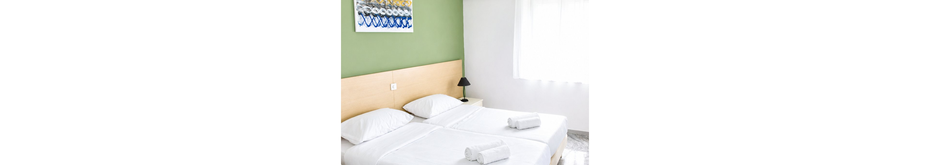 A clean, minimalist bedroom features two neatly made twin beds with white linens and rolled towels placed on top. The room has a light wooden headboard, a small bedside table with a black lamp, and a green accent wall displaying abstract artwork. Sunlight filters through sheer curtains covering a window, illuminating the polished stone floor. The overall atmosphere is bright, modern, and inviting, suggesting a hotel or guest room setting.