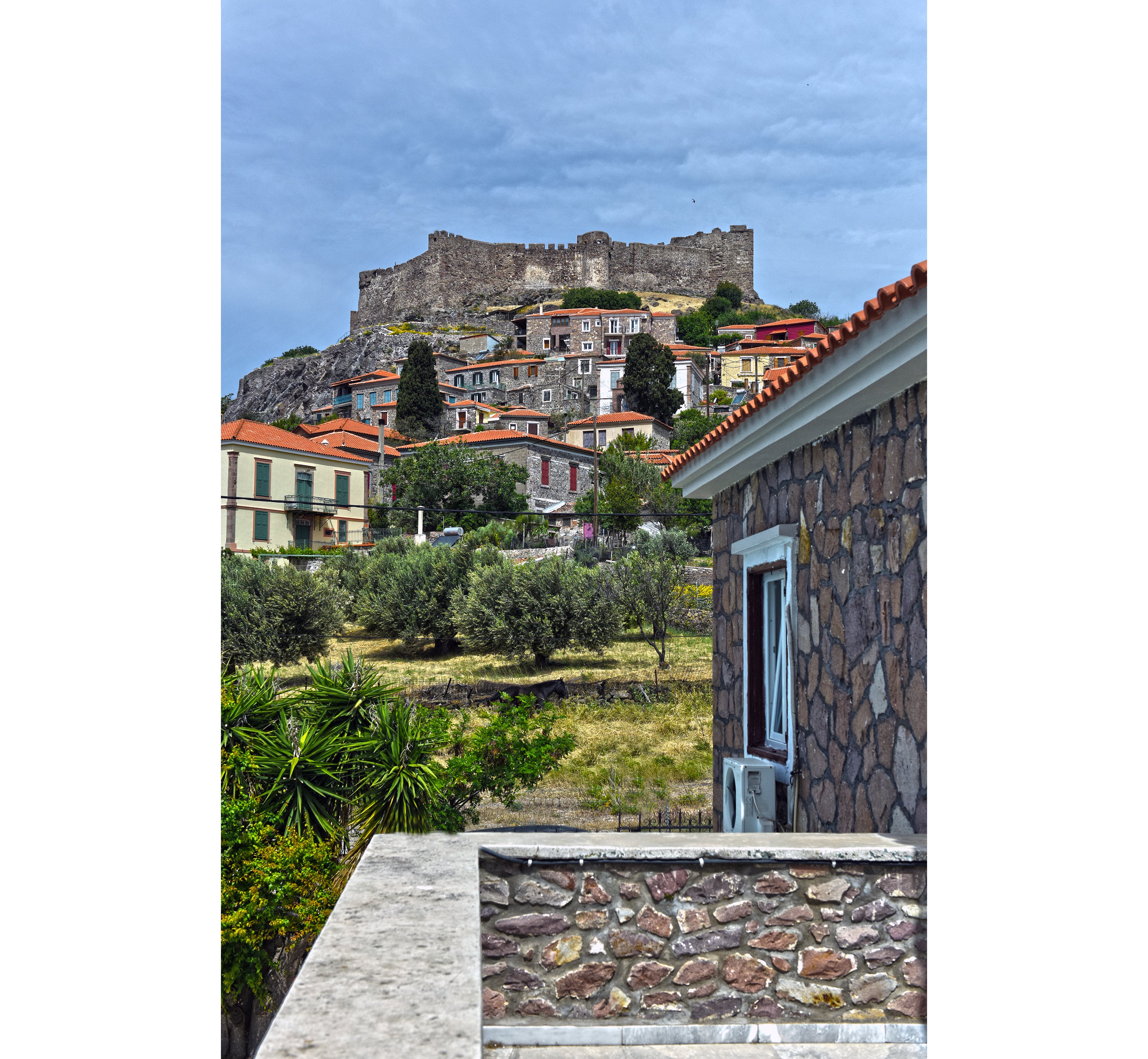 A historic stone village climbs a hillside beneath an ancient fortress. Traditional houses with red-tiled roofs are surrounded by trees and greenery. In the foreground, a stone-walled building and terrace frame the view toward the castle. The sky is overcast, adding a dramatic contrast to the earthy tones of the architecture. The scene evokes a quiet, old-world Mediterranean atmosphere.