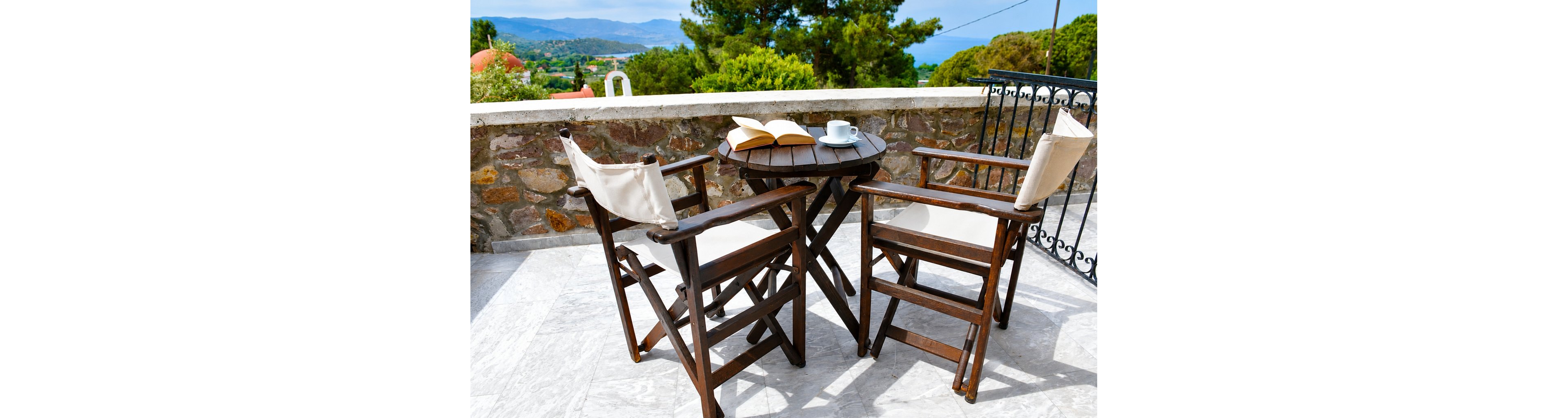 A small round wooden table with two wooden chairs sits on a bright stone terrace. An open book and a cup of coffee rest on the table, suggesting a quiet reading moment. The terrace overlooks a scenic landscape with trees, hills, and distant water. Warm sunlight and clear skies create a relaxed, inviting atmosphere.