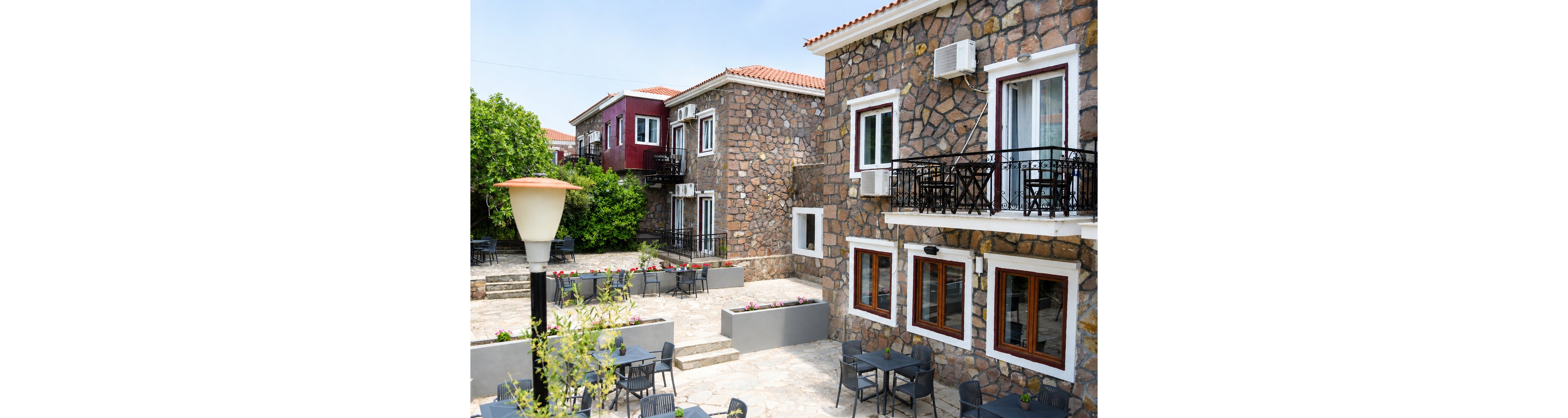 A charming stone courtyard is surrounded by rustic multi-story buildings with red-tiled roofs. Several black tables and chairs are arranged on the terrace, suggesting an outdoor cafe or dining area. Flower boxes with colorful plants line the low walls, adding greenery to the scene. A large lamp post and balconies with metal railings contribute to the relaxed, Mediterranean-style atmosphere.