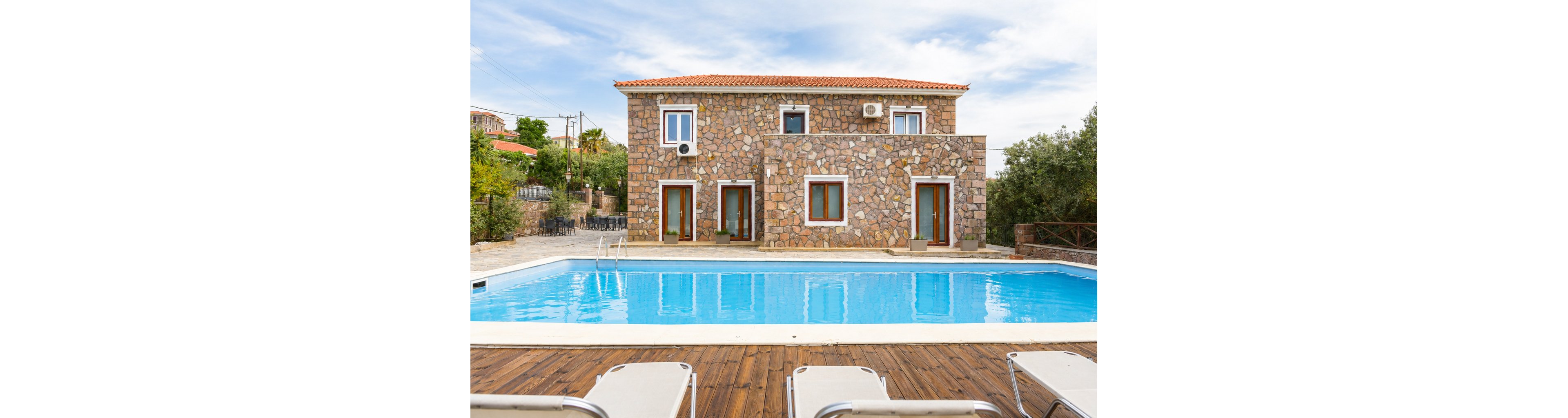 A two-story stone villa stands in front of a clear blue swimming pool. The scene features wooden decking with sun loungers in the foreground, creating a relaxing resort atmosphere. Multiple windows and doors face the pool, along with visible air conditioning units on the facade. Trees and other buildings appear in the background under a partly cloudy sky, suggesting a peaceful vacation setting.