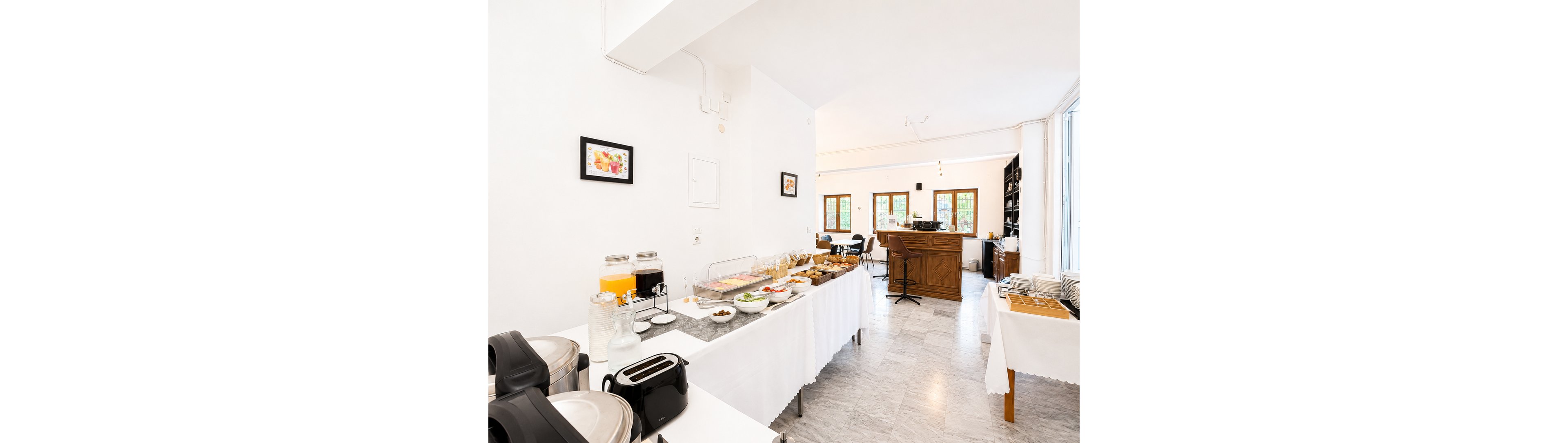 A bright self-service breakfast buffet is arranged on long white-covered tables in a modern dining room. Various foods, cereals, and pastries are displayed alongside glass dispensers of juice and water. Toasters, chafing dishes, and other breakfast appliances are set up for guests. In the background, a wooden bar counter and large windows create an inviting, airy atmosphere.
