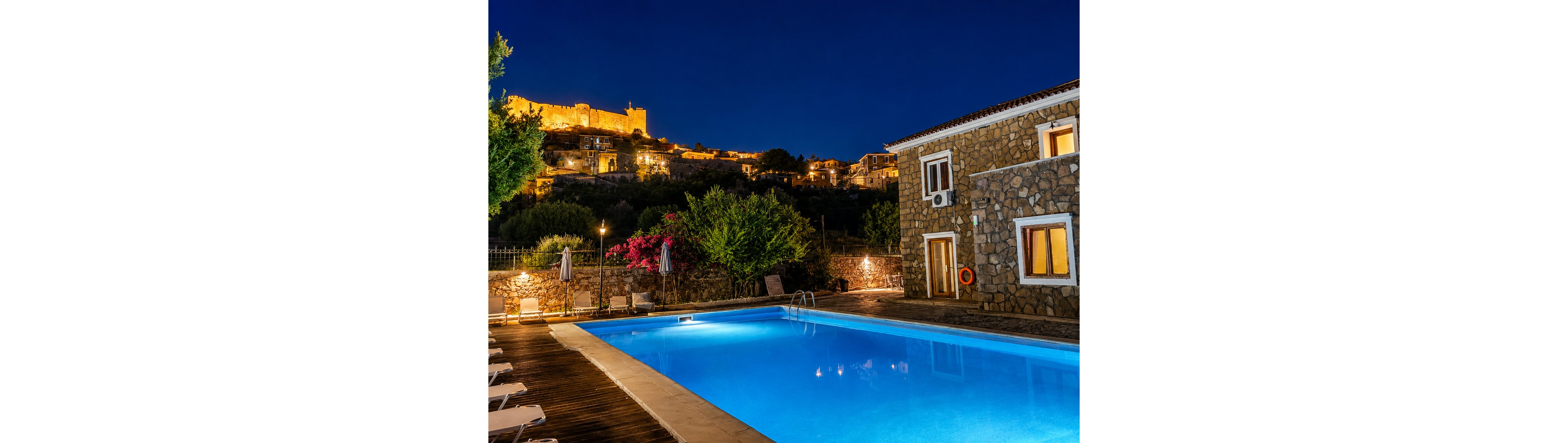A brightly lit swimming pool glows blue at night beside a stone building. Lounge chairs and closed umbrellas line the wooden deck around the pool. In the background, an illuminated hilltop fortress and village walls stand out against the deep blue sky. Warm yellow lights contrast with the cool tones of the water, creating a tranquil evening atmosphere.