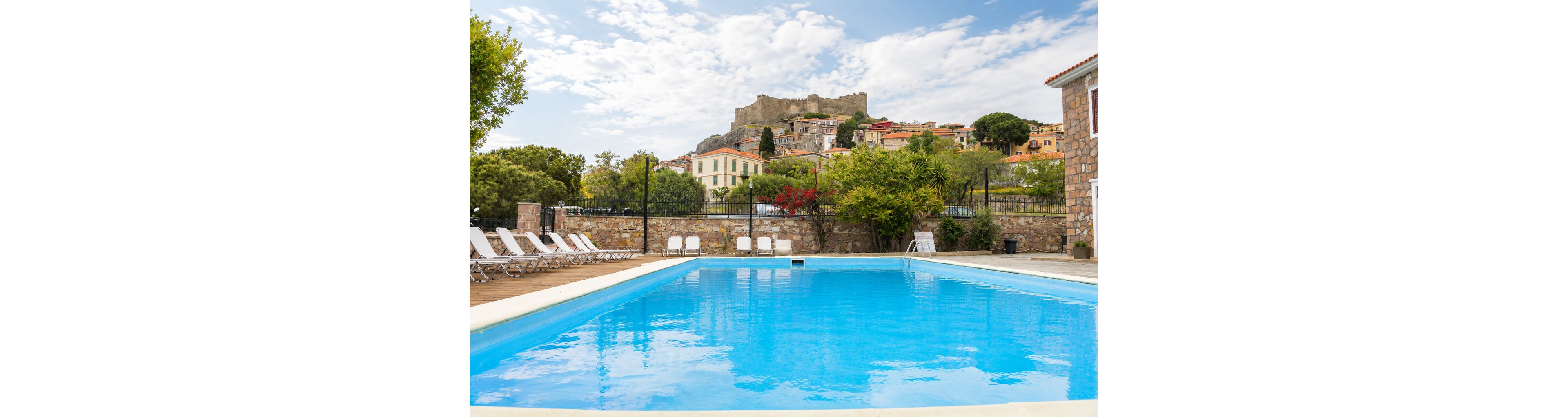 A clear blue swimming pool sits in the foreground of a sunny resort courtyard. White lounge chairs line the pool deck, facing the water and a black metal fence. Beyond the pool, colorful houses and lush greenery climb a gentle hillside. At the top of the hill, an old stone fortress overlooks the village under a partly cloudy sky. The scene feels calm and inviting, ideal for a relaxing vacation setting.