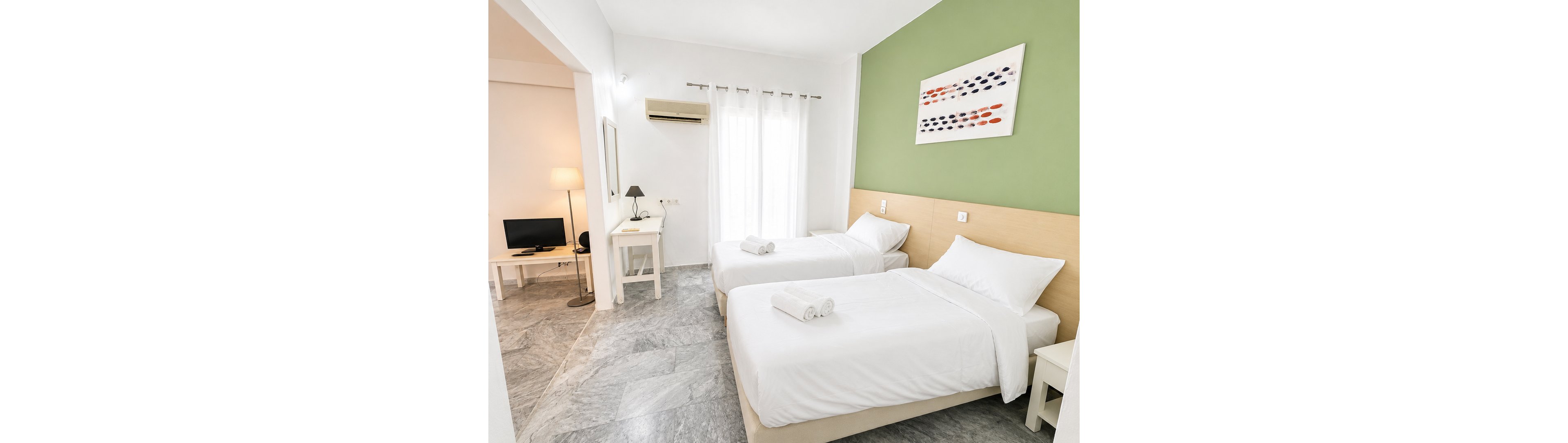 Contemporary hotel room featuring two neatly made twin beds with white linens and folded towels. The space is bright, with natural light coming through sheer curtains and a green accent wall behind the beds. A small desk, chair, and wall-mounted air conditioner are visible, along with a separate sitting area containing a TV and floor lamp. Clean marble-style flooring and minimalist decor create a fresh, welcoming atmosphere.
