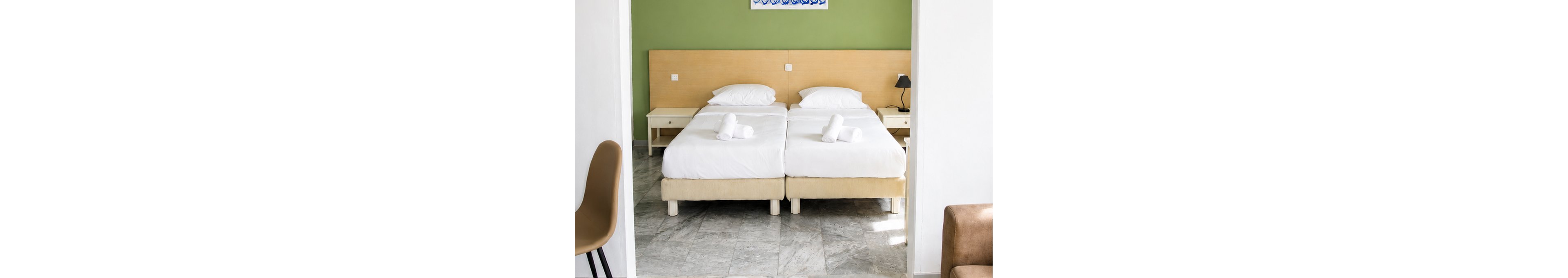 A bright, minimalist hotel room features two neatly made twin beds with white linens and rolled towels placed on top. The beds sit against a light wooden headboard and a green accent wall, with a small bedside table and lamp to one side. In the foreground, a simple coffee table with a small plant and modern chairs frame the view into the sleeping area. Natural light enhances the clean, contemporary design and neutral color palette.
