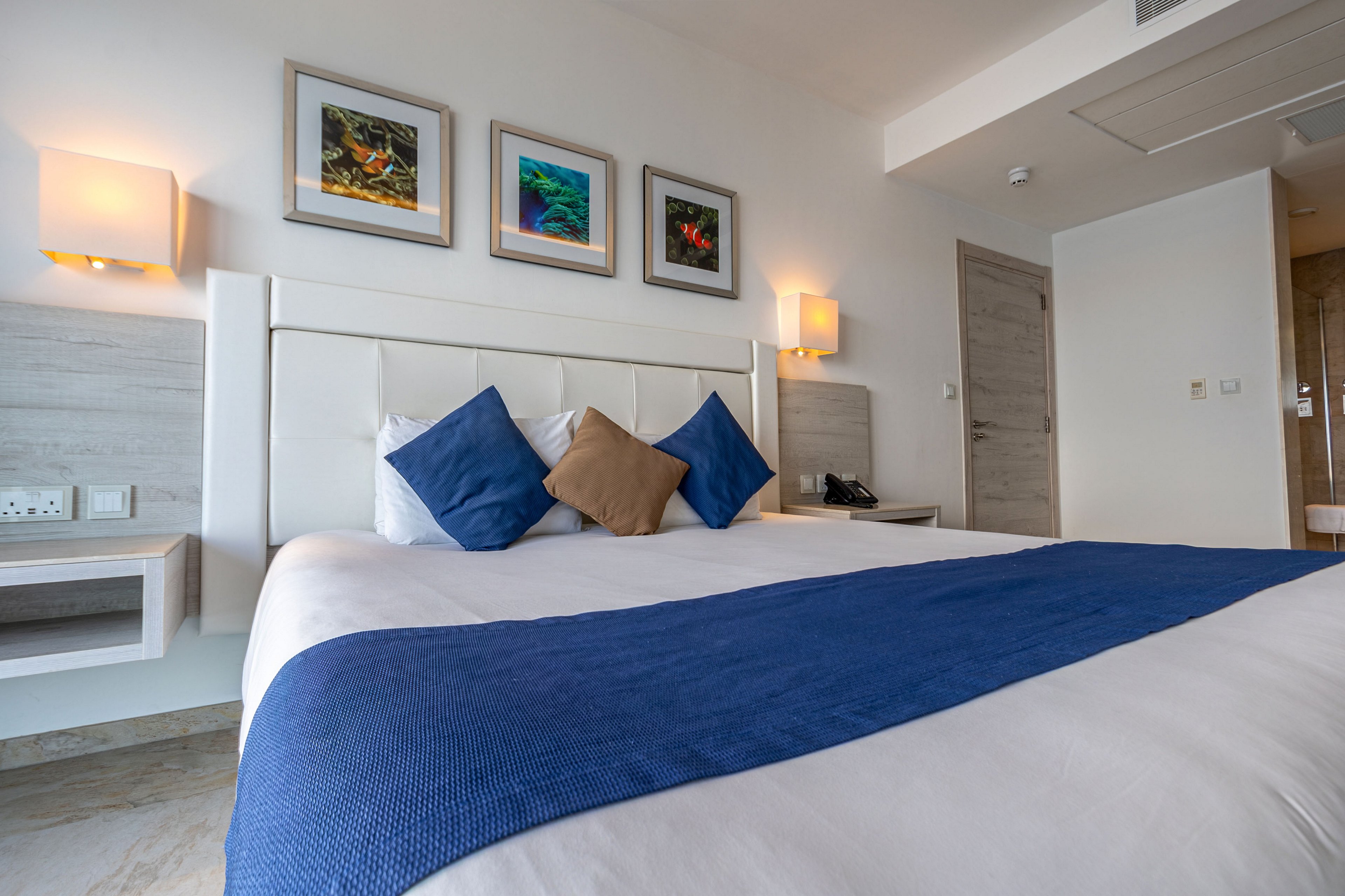 Contemporary hotel room interior featuring a large bed with white linens and a blue runner. Three decorative pillows in blue and brown are arranged against a padded white headboard. Wall-mounted lamps and framed artwork add warmth and color to the minimalist decor. A bedside table, telephone, and partially visible bathroom suggest a comfortable guest accommodation setting.