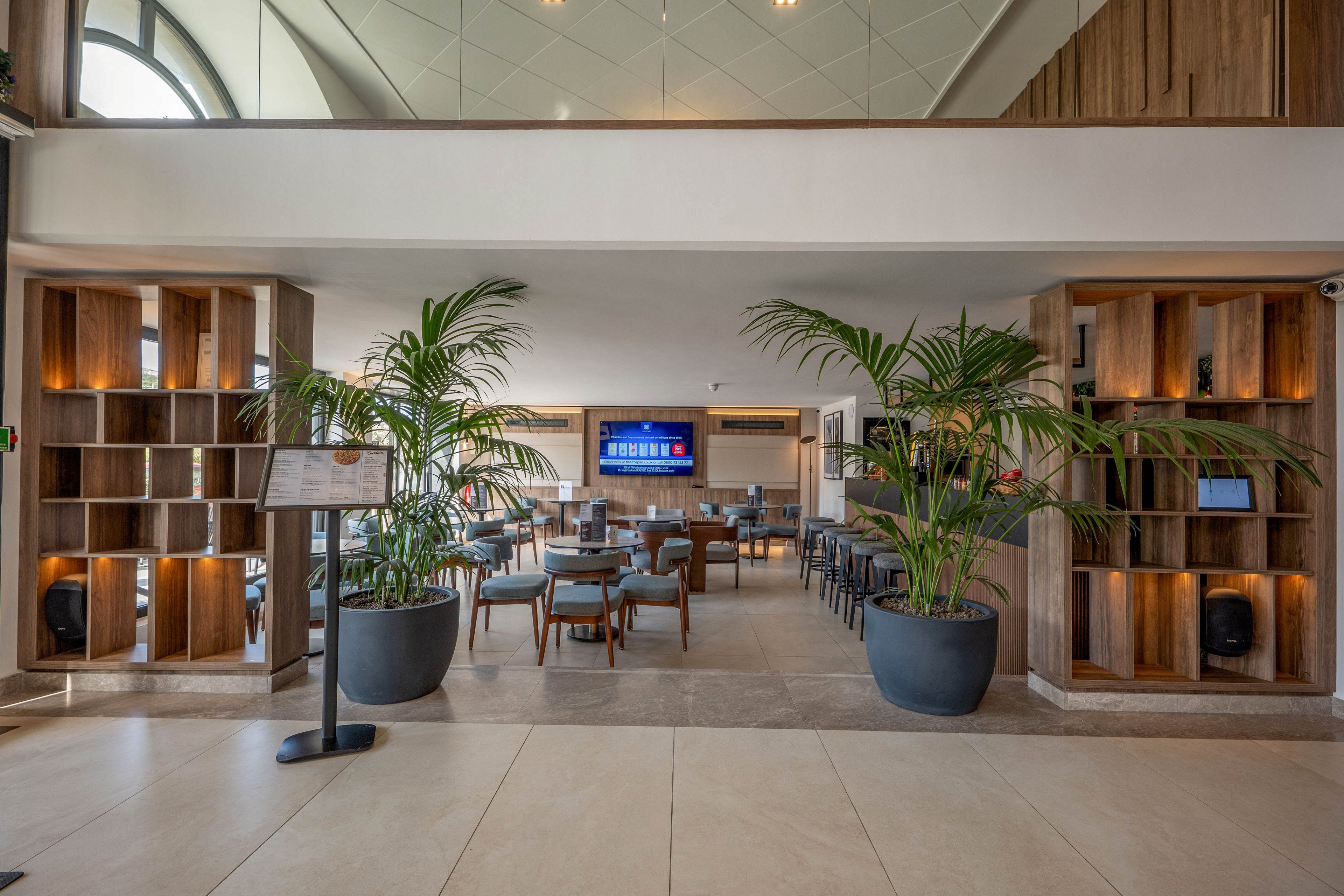 A contemporary hotel lounge features neatly arranged tables and chairs in a spacious open-plan setting. Large potted plants frame the entrance, adding greenery to the neutral-toned interior. Wooden shelving units with decorative objects line both sides, while a digital screen is mounted on the far wall. Soft natural light enters through high windows, creating a relaxed and welcoming atmosphere.