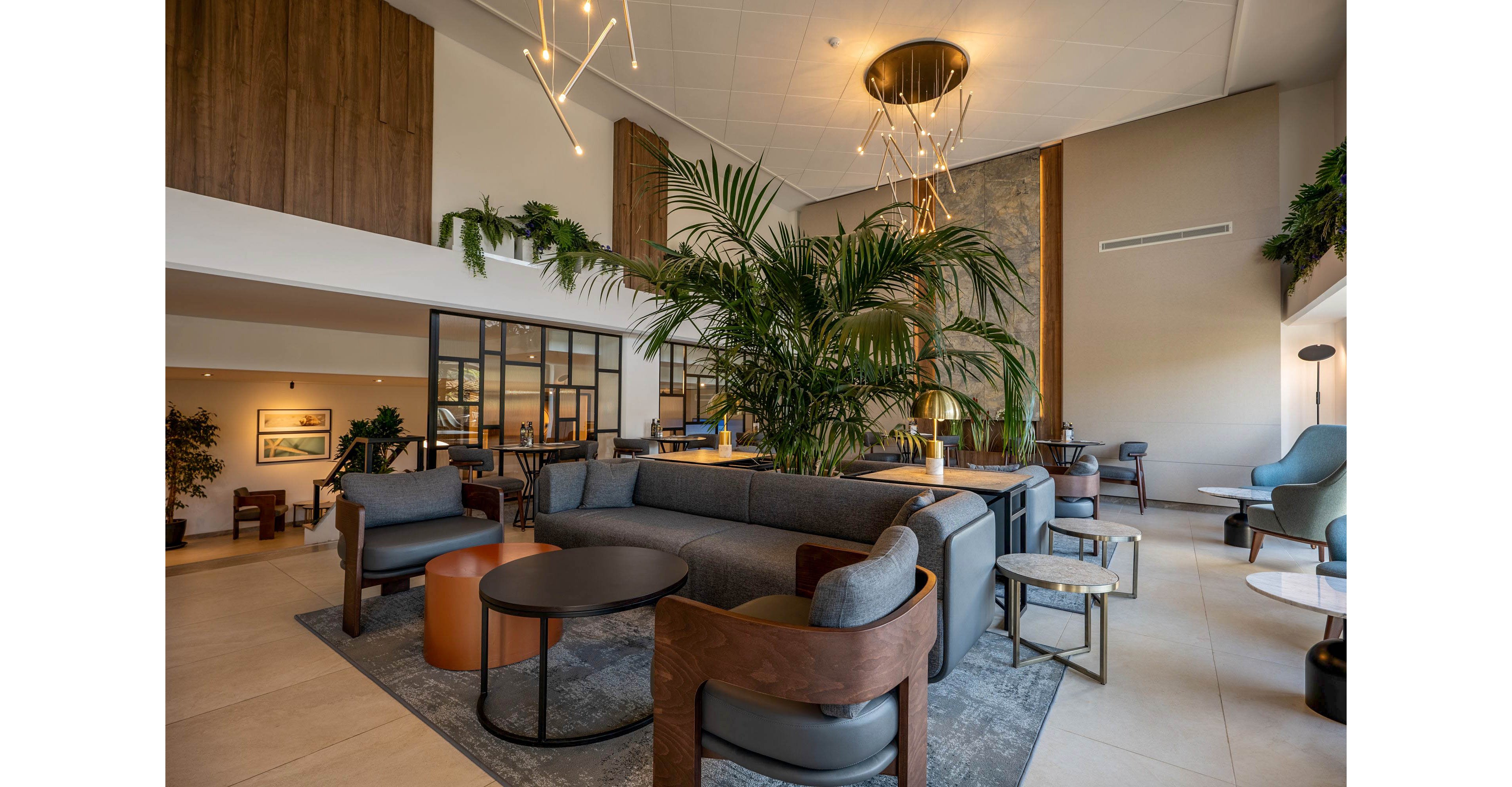 A spacious modern lobby features contemporary lounge seating arranged around low circular tables. Large indoor plants create a lush focal point in the center of the room. Warm wood accents, neutral upholstery, and minimalist lighting fixtures contribute to a sophisticated atmosphere. Floor-to-ceiling windows and glass partitions add openness and natural light to the interior.