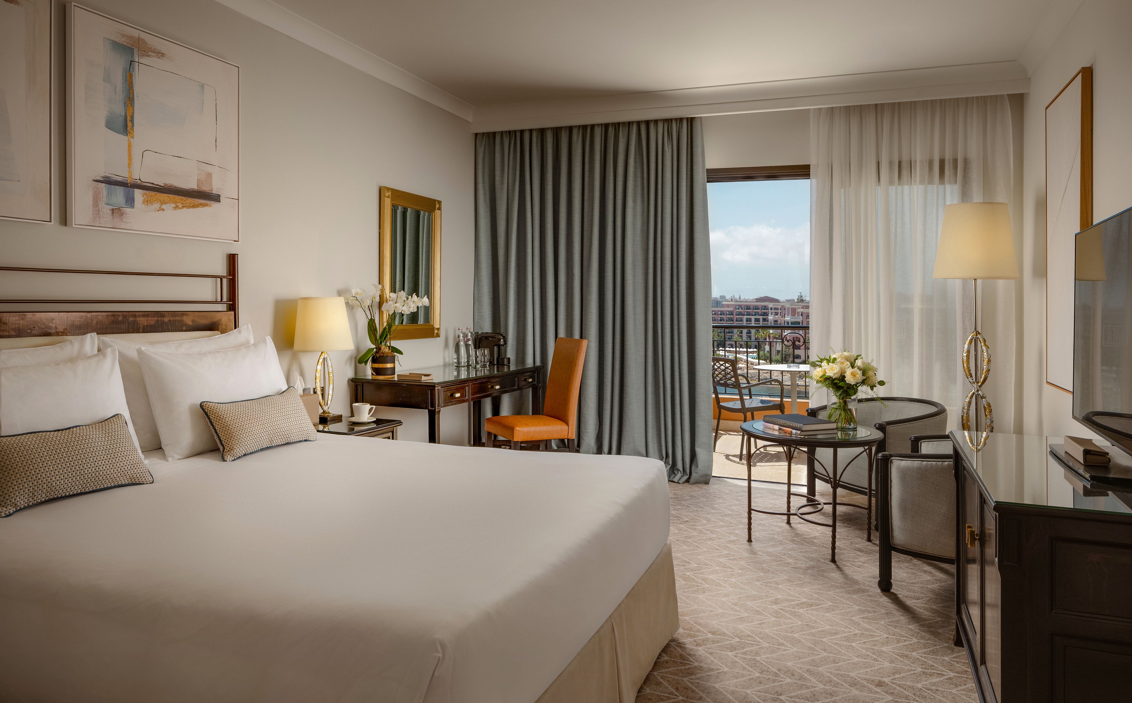 A spacious, well-lit hotel room features a large neatly made bed with white linens and decorative pillows. The room includes a wooden desk with an orange chair, a floor lamp, and a small round table with chairs near the balcony. Neutral tones and soft lighting create a calm, sophisticated atmosphere, complemented by framed artwork and fresh flowers. Sheer curtains reveal a balcony overlooking an urban or waterfront landscape under a bright sky.