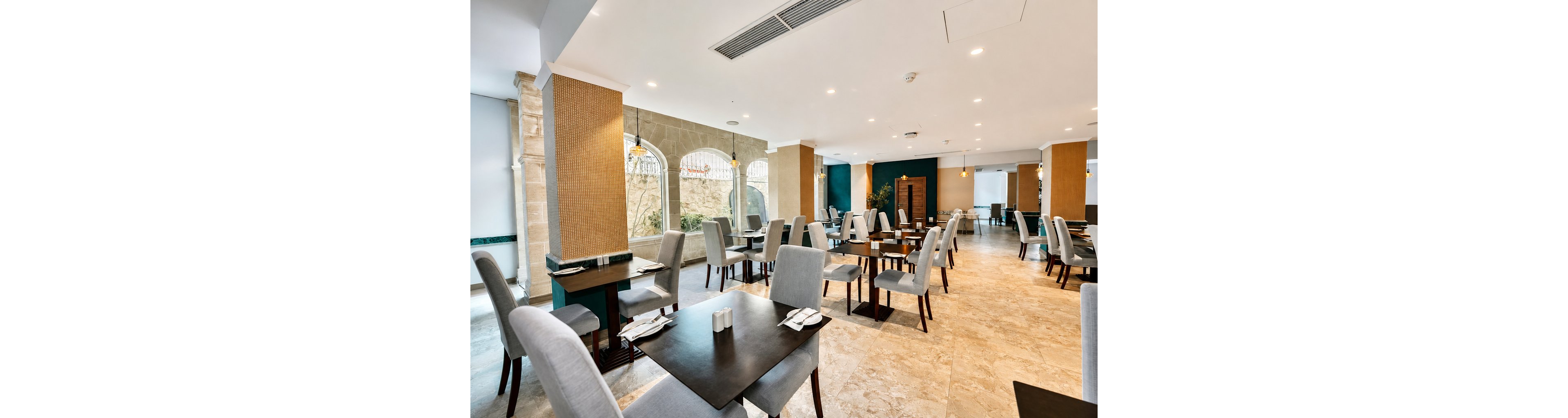 A contemporary restaurant dining area features neatly arranged dark wooden tables and light gray upholstered chairs. The space is empty of people, highlighting a clean and orderly setup with place settings and napkins prepared. Large arched windows along one wall allow natural light to brighten the room, complemented by recessed ceiling lights and warm accent lighting. Neutral tones, stone flooring, and minimalist decor create a calm, upscale atmosphere suitable for hotel or business dining.