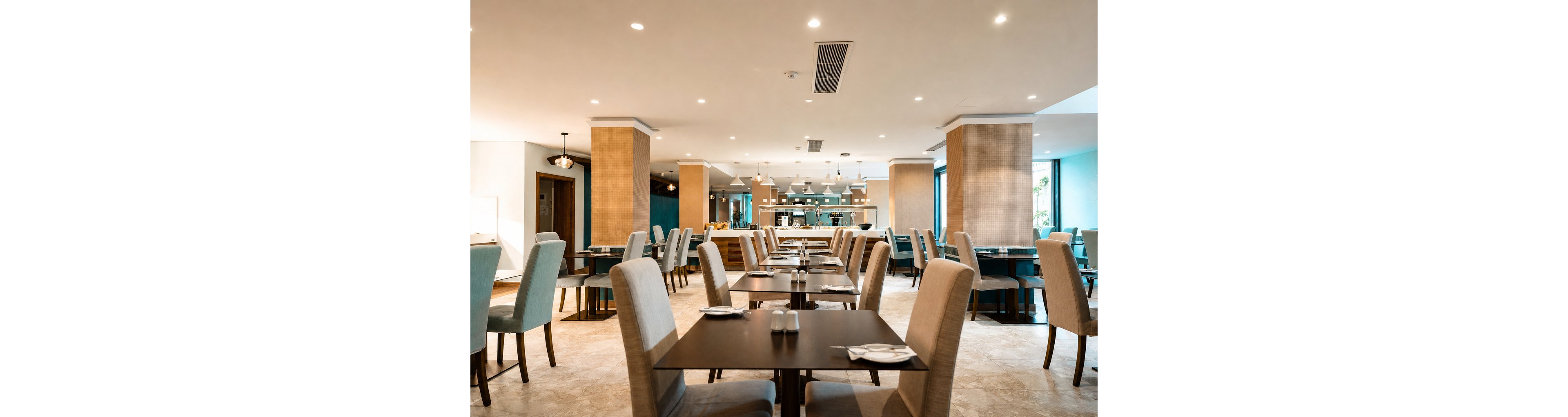A contemporary restaurant dining room features neatly arranged tables and upholstered chairs in soft blue and beige tones. The space is empty, with place settings prepared on dark wooden tables under warm ceiling lights. Large columns divide the room, leading toward an open kitchen or serving area at the back. Natural light enters through tall windows, enhancing the clean and inviting atmosphere.