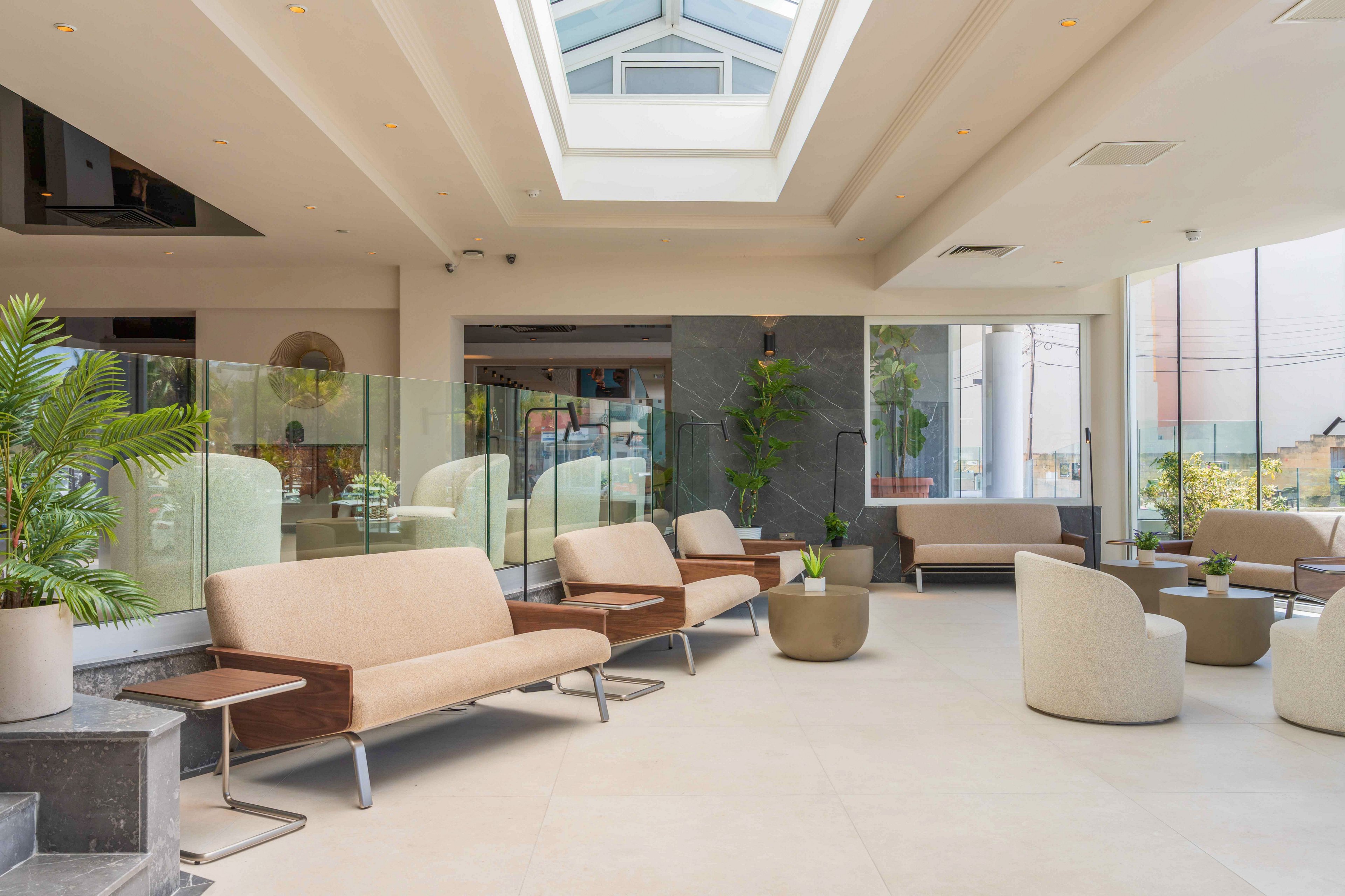 A spacious contemporary lobby features light beige sofas and armchairs arranged along glass partitions and low tables. Large windows and a central skylight flood the interior with natural daylight, highlighting the clean architectural lines. Potted green plants add a touch of nature and contrast against the neutral flooring and walls. The overall atmosphere is calm, inviting, and suitable for waiting or informal meetings.