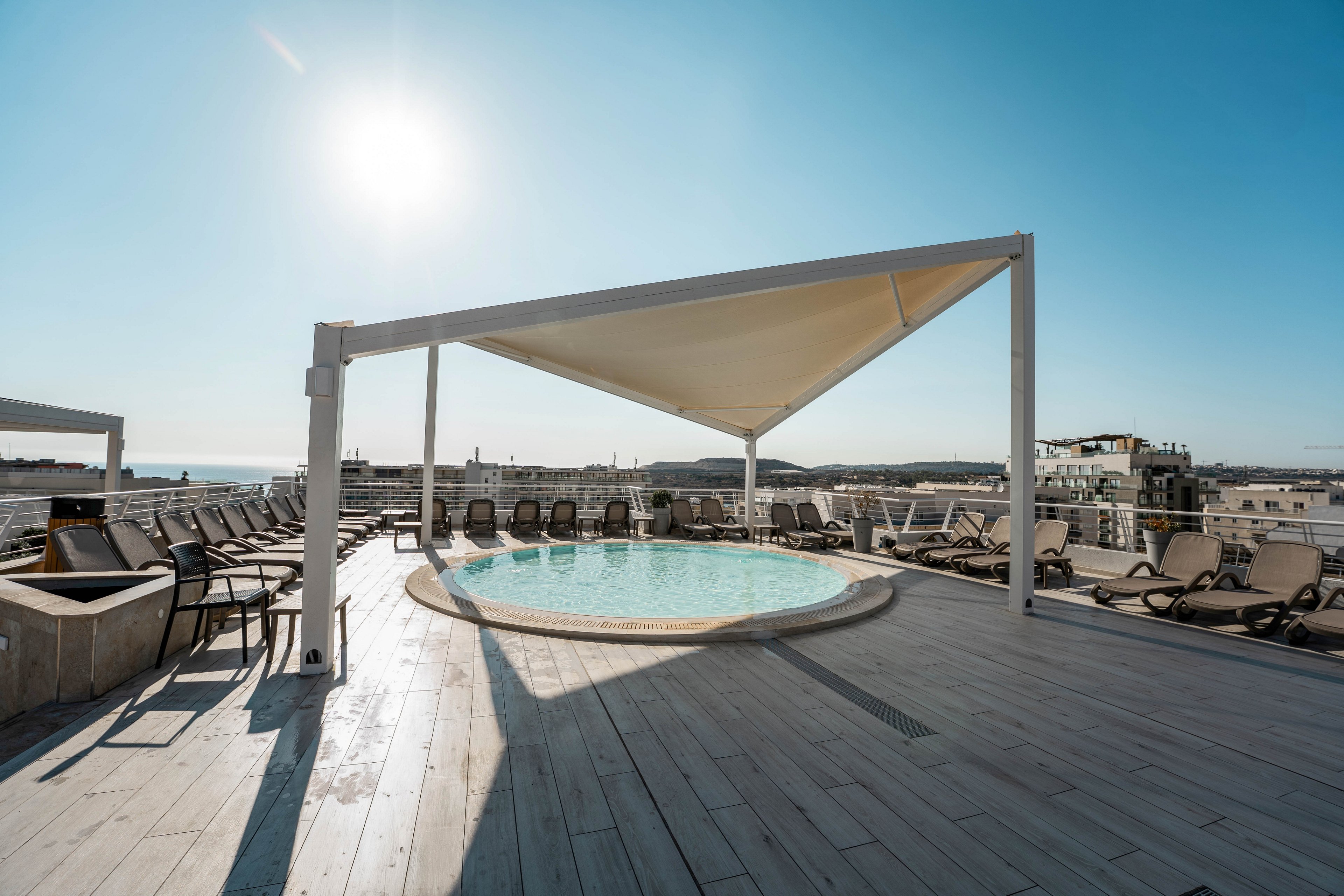 A circular rooftop pool sits beneath a modern triangular shade canopy on a spacious wooden deck. Numerous sun loungers line the perimeter, facing outward toward the surrounding cityscape and distant coastline. The bright sun and clear blue sky create a warm, inviting atmosphere ideal for relaxation. The scene suggests a contemporary hotel or apartment terrace designed for leisure and sunbathing.