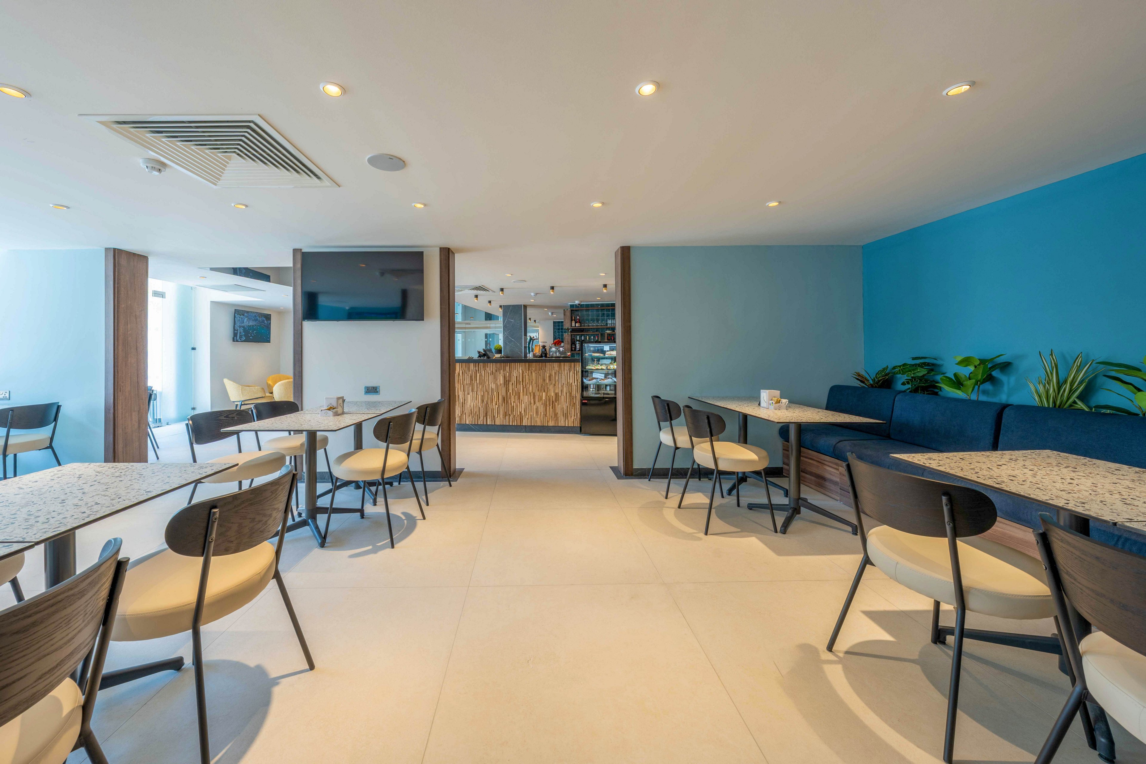 Bright contemporary café interior featuring multiple tables and chairs arranged in an open layout. A blue accent wall with built-in bench seating and plants adds color to the neutral space. Recessed ceiling lights and large floor tiles create a clean, minimalist atmosphere. The counter area in the background suggests a relaxed dining or coffee shop setting.