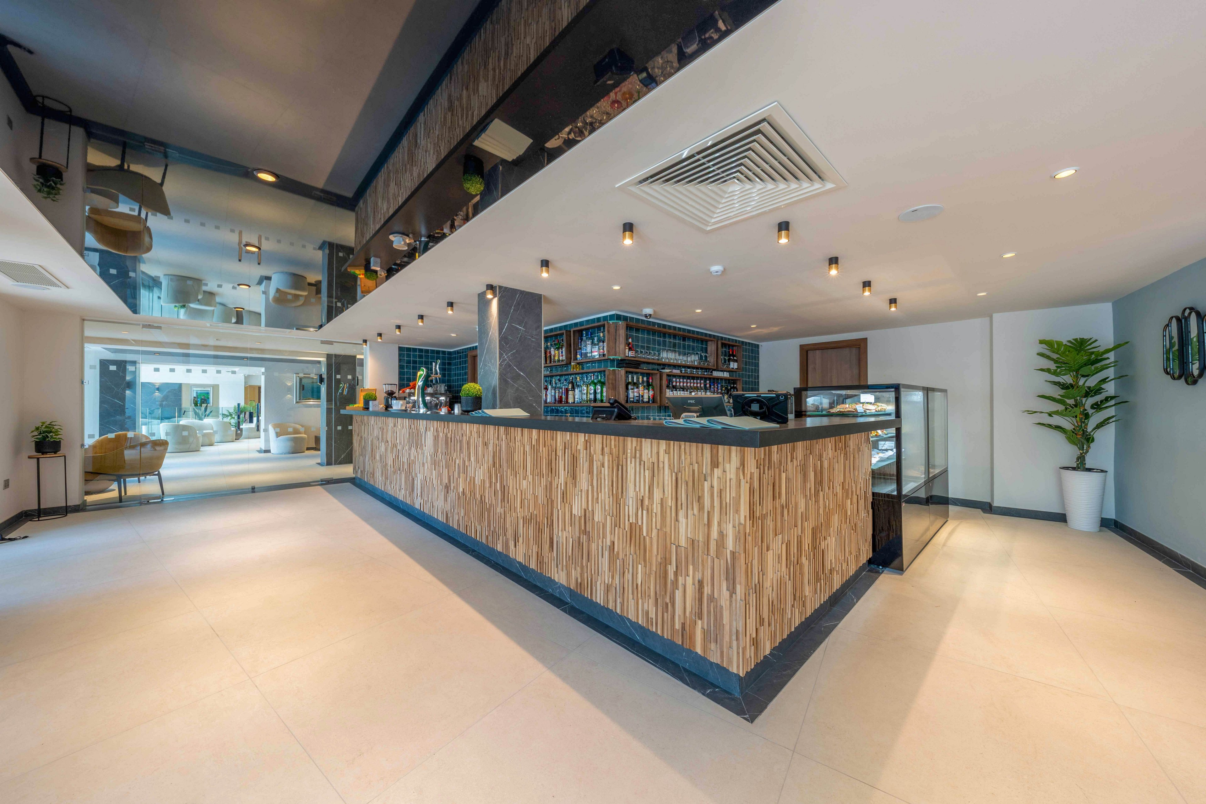A spacious contemporary lobby features a large wooden bar counter as the central focal point. The bar area includes shelves with bottles and glassware, and a glass display case on one side. Recessed ceiling lights and hanging fixtures provide warm illumination across the tiled floor. A potted plant and lounge seating in the background add a welcoming, relaxed atmosphere.