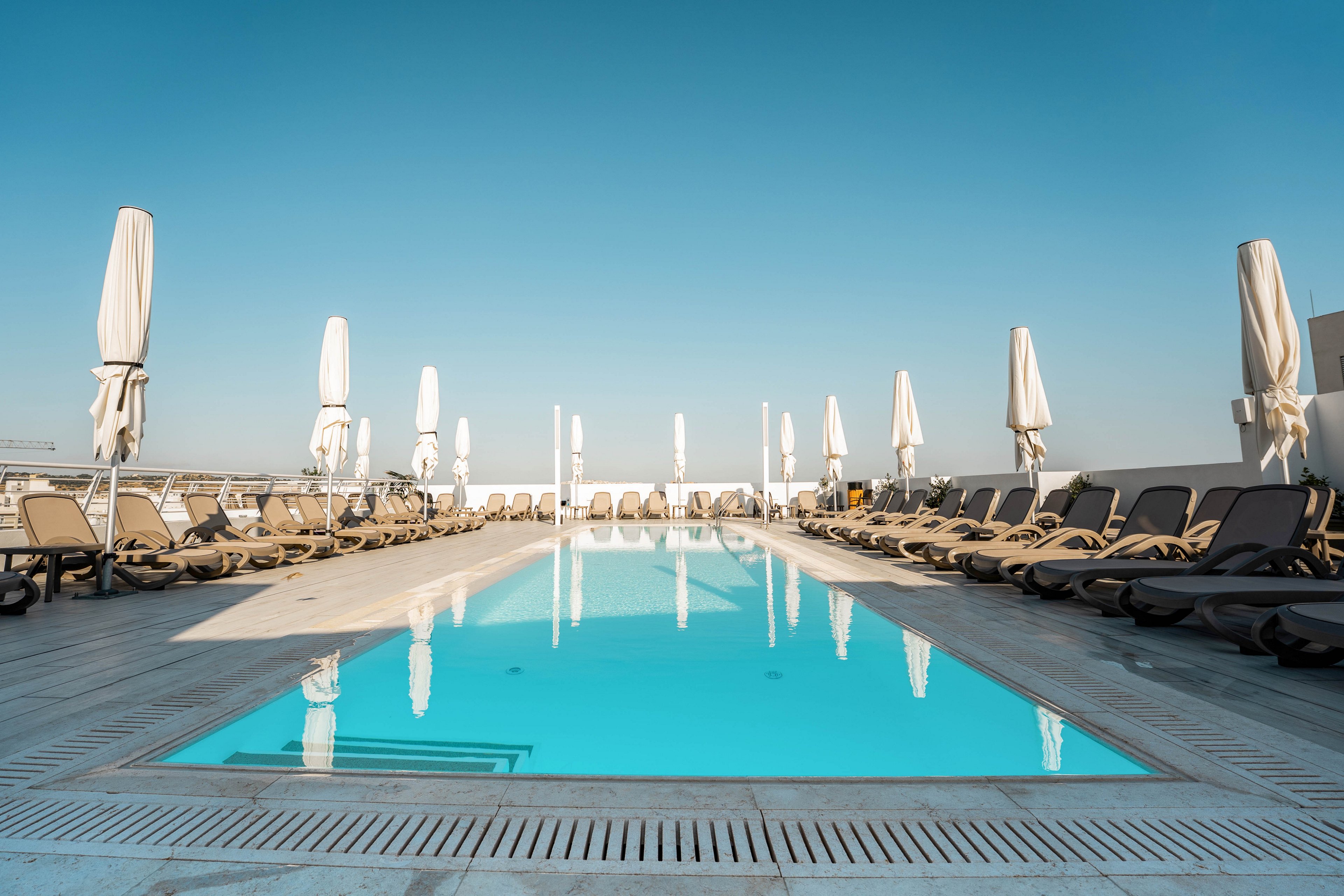 A clear blue swimming pool sits at the center of a modern rooftop terrace. Rows of empty sun loungers line both sides of the pool, each paired with a closed white umbrella. The sky is cloudless and bright, emphasizing the tranquil and minimalist atmosphere. The scene suggests a quiet resort or hotel setting before guests arrive.