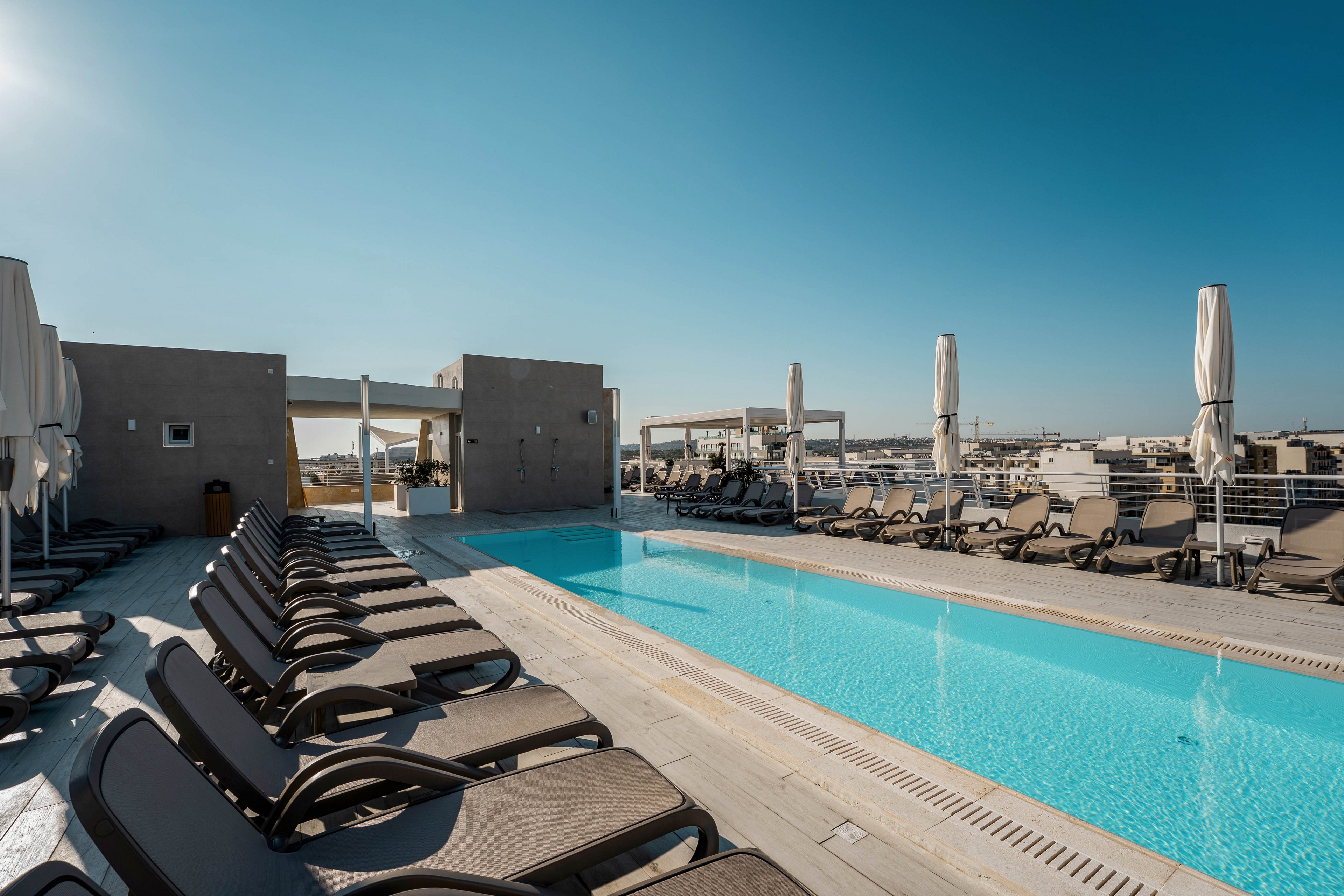 A sleek rooftop swimming pool is lined with rows of brown sun loungers under a clear blue sky. Closed white umbrellas stand between the loungers, suggesting a sunny, warm day. The pool’s turquoise water contrasts with the light stone deck and minimalist architectural structures. In the distance, a cityscape stretches toward the horizon, emphasizing an urban, resort-like atmosphere.