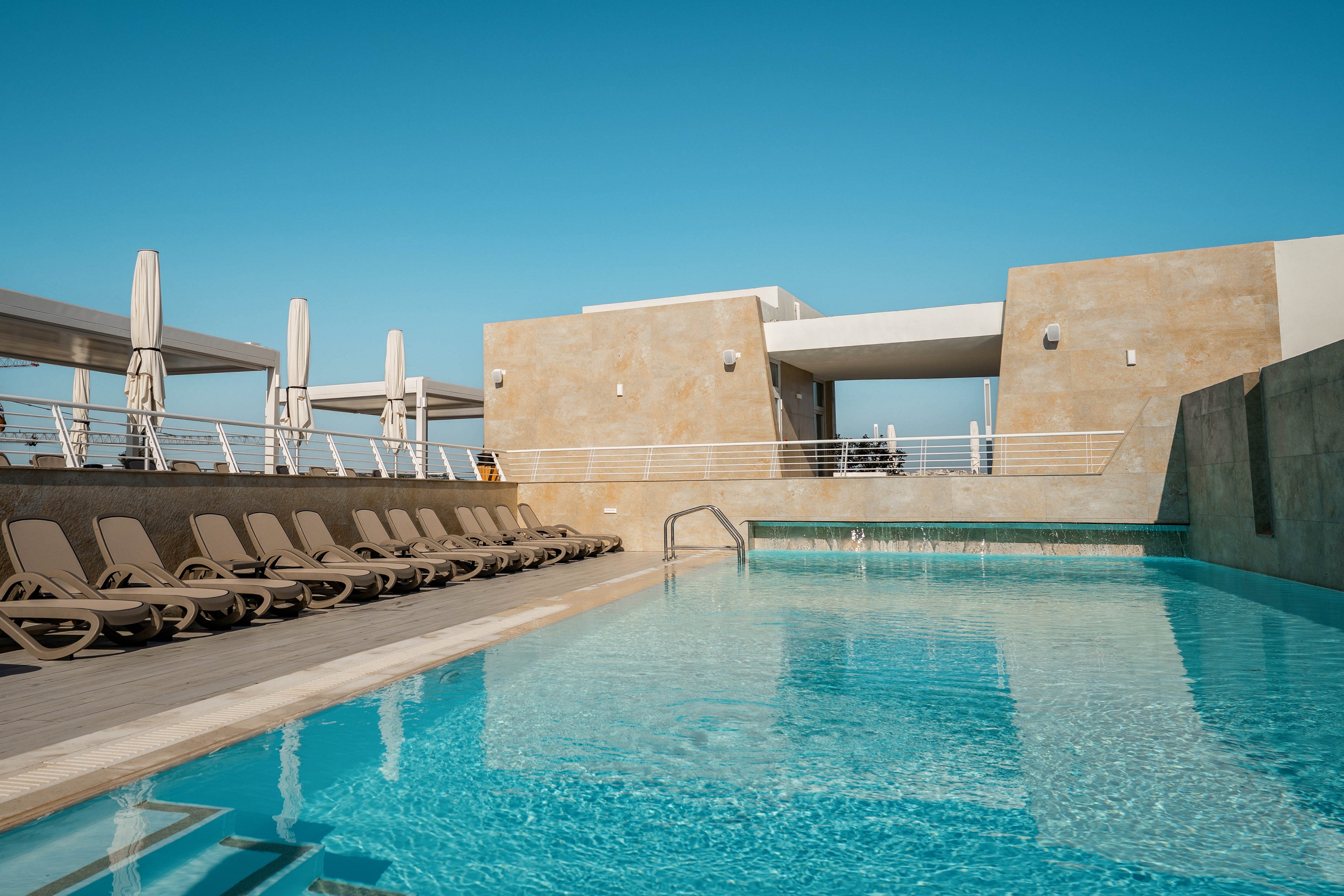 A clear blue swimming pool sits on a modern rooftop terrace under a bright sky. Rows of empty sun loungers line one side of the pool, facing contemporary concrete and white structures. Tall closed umbrellas stand behind the loungers, suggesting a resort or hotel setting. The scene feels calm and minimalistic, emphasizing clean lines, water reflections, and warm sunlight.