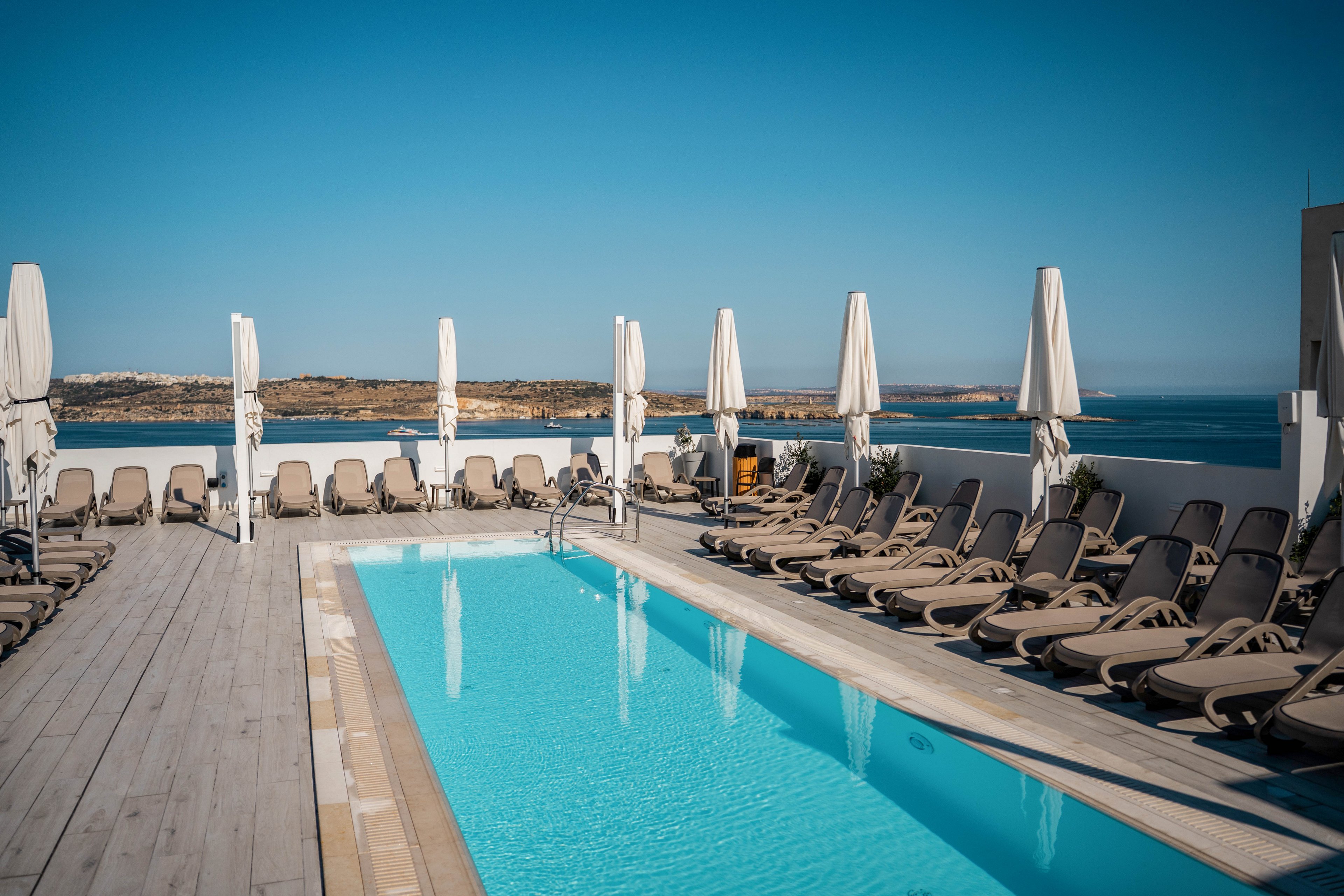 An outdoor rooftop swimming pool is surrounded by rows of empty sun loungers and closed white umbrellas. The scene overlooks a calm blue sea and distant coastline under a clear sky. Wooden decking frames the pool area, creating a clean and modern resort atmosphere. The bright sunlight and vivid blue water suggest a warm, relaxing vacation setting.
