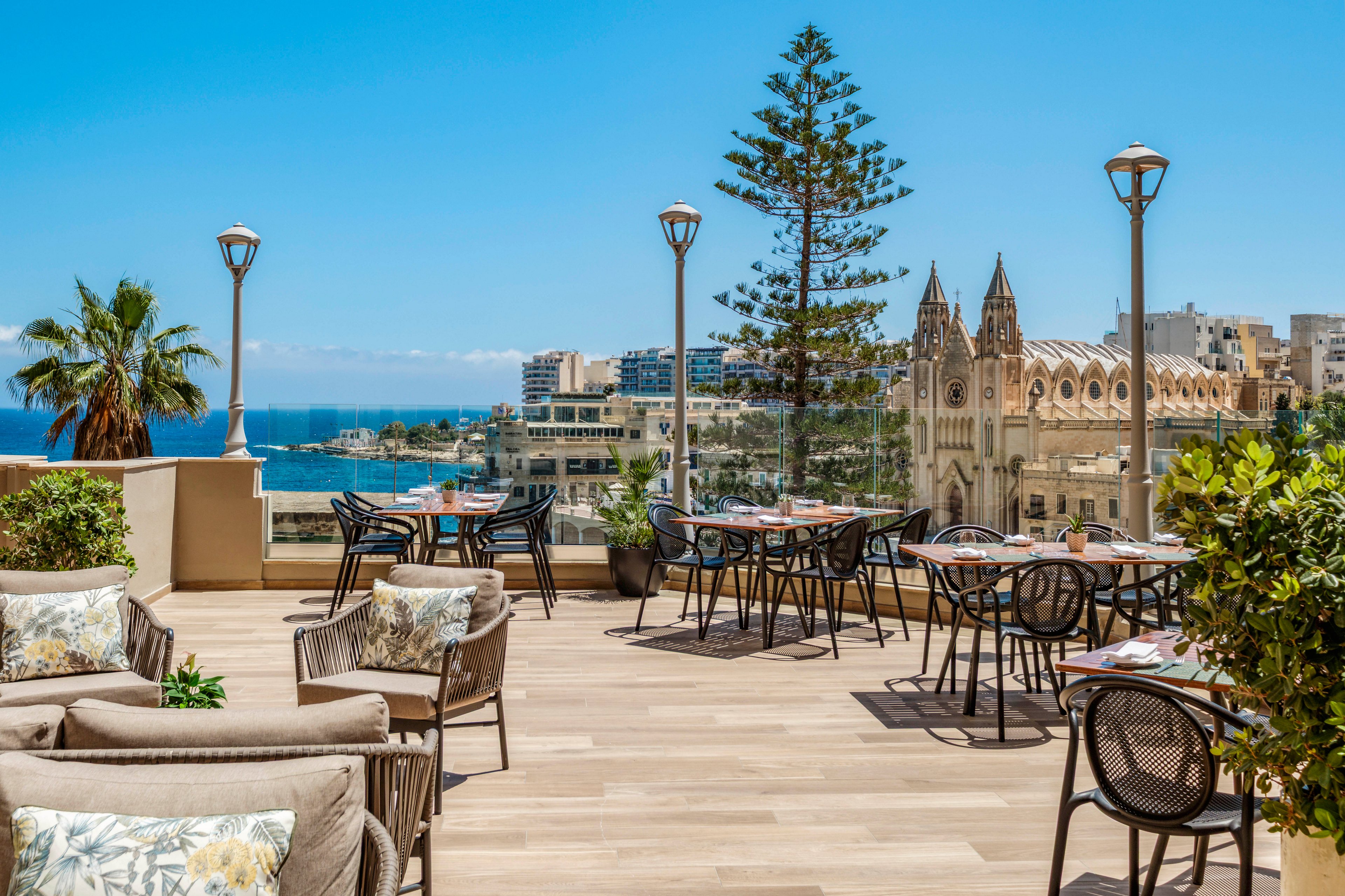 An inviting rooftop terrace features outdoor tables and cushioned lounge chairs arranged on a wooden deck. The setting overlooks a coastal cityscape with historic architecture, tall trees, and the sea in the background under a clear blue sky. Potted plants and greenery frame the seating area, creating a relaxed and stylish atmosphere. The scene suggests a tranquil dining or social space with scenic ocean views.