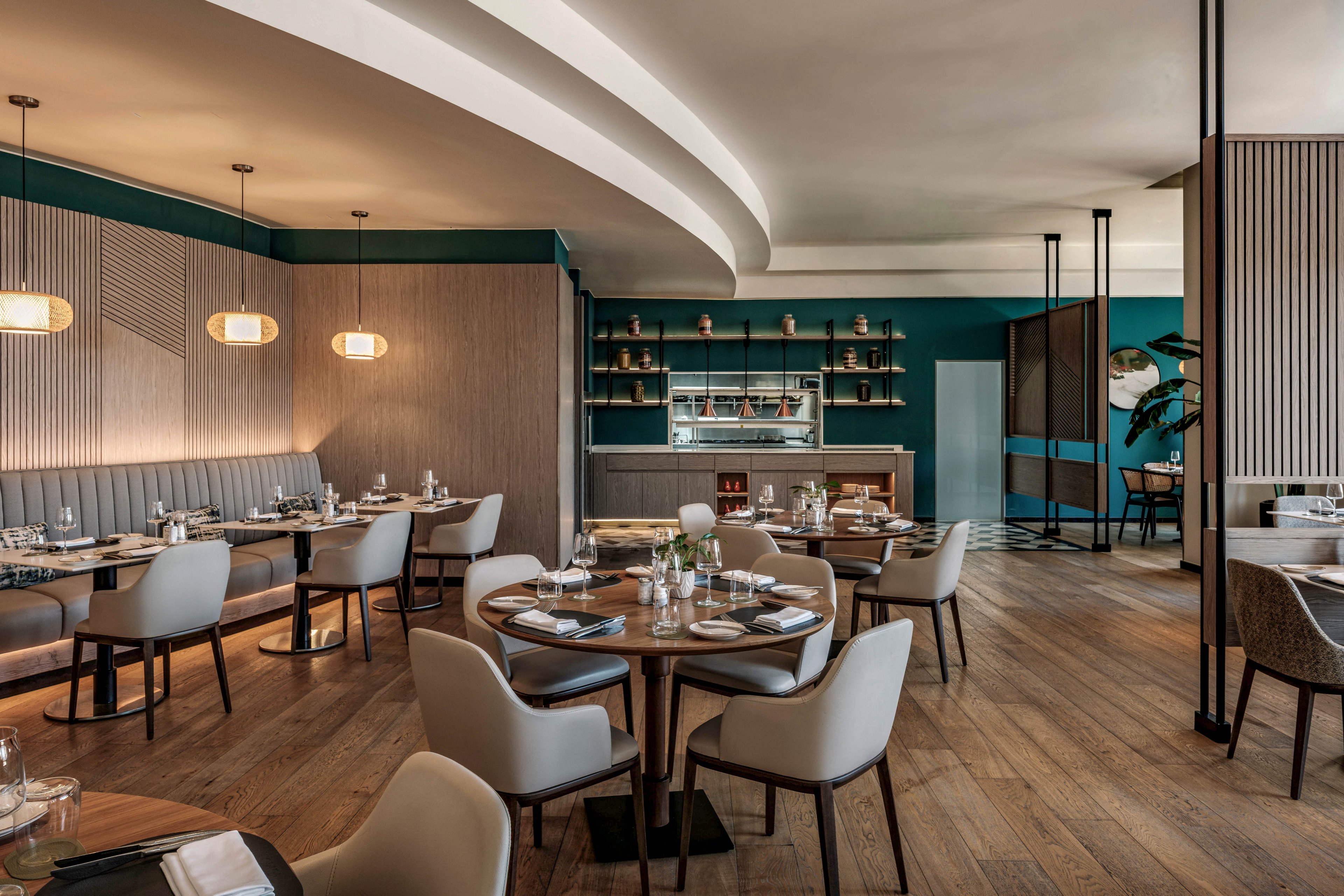 A modern restaurant dining room features round wooden tables set with glassware and plates. Soft upholstered chairs surround the tables, and built-in banquette seating lines the wall. Warm wood flooring contrasts with teal accent walls and minimalist pendant lights. Shelving and an open kitchen area are visible in the background, creating a stylish yet inviting atmosphere.