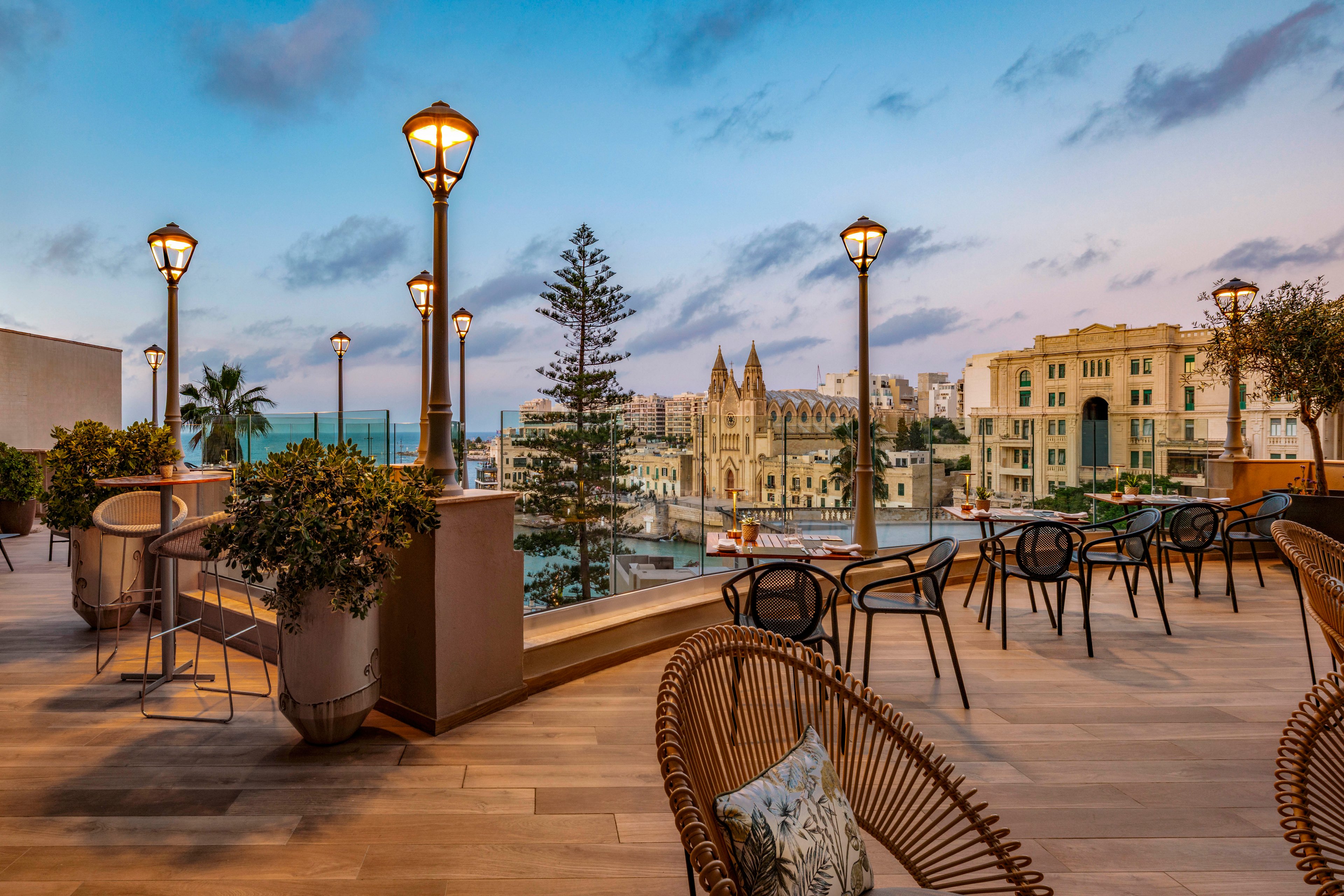 An upscale outdoor terrace overlooks a tranquil waterfront and historic church at dusk. Warm streetlights illuminate modern chairs, tables, and potted plants on the wooden deck. The sky glows with soft pastel colors as evening settles over the surrounding buildings and calm water. The scene evokes a relaxed, sophisticated atmosphere ideal for dining or socializing.