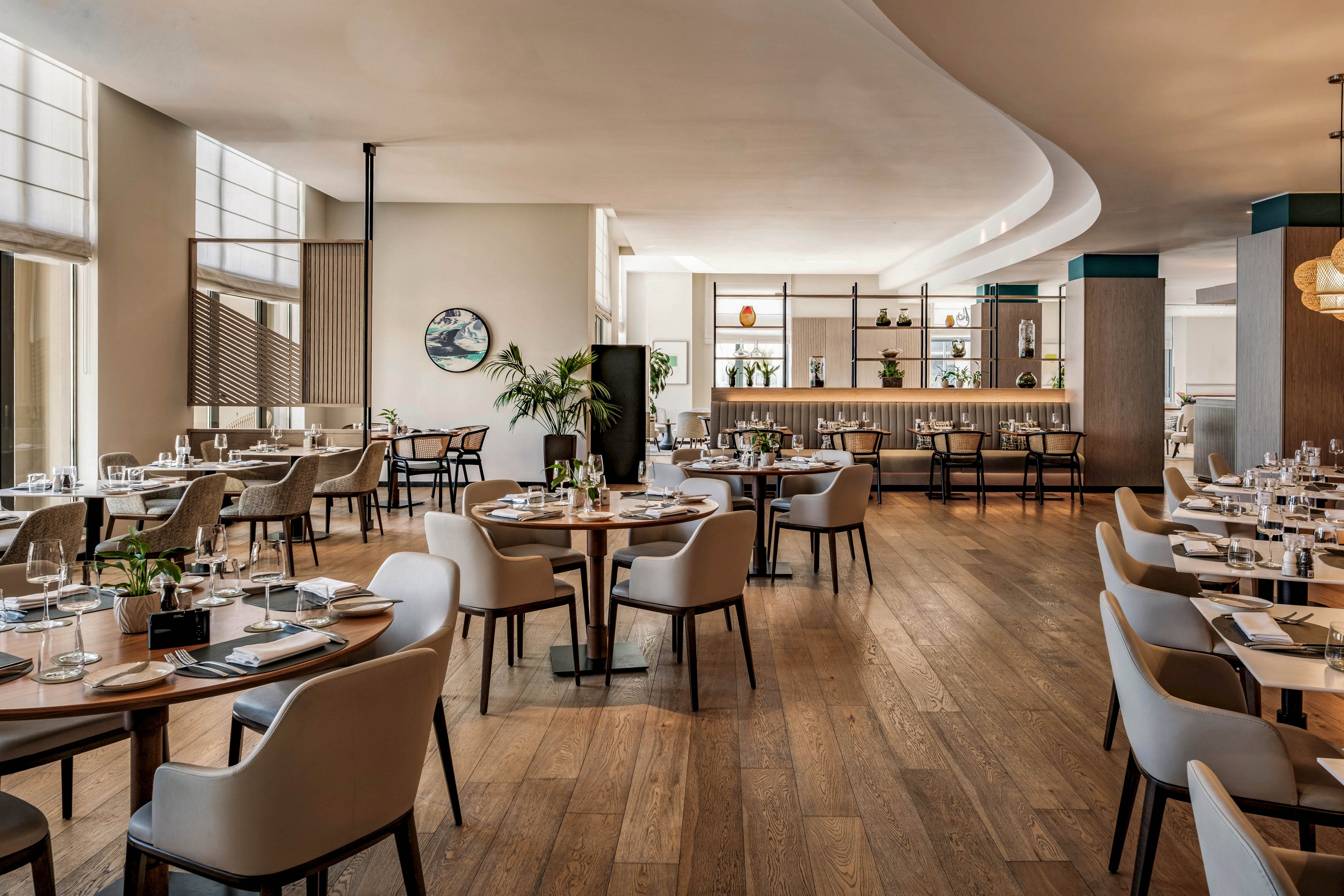 Contemporary restaurant dining room featuring round wooden tables and upholstered chairs is shown in a bright, spacious setting. Large windows allow natural light to fill the space, highlighting the wood flooring and minimalist decor. A stylish bar area with shelves of bottles and plants appears in the background, adding depth and visual interest. The overall atmosphere feels elegant, welcoming, and designed for fine dining.