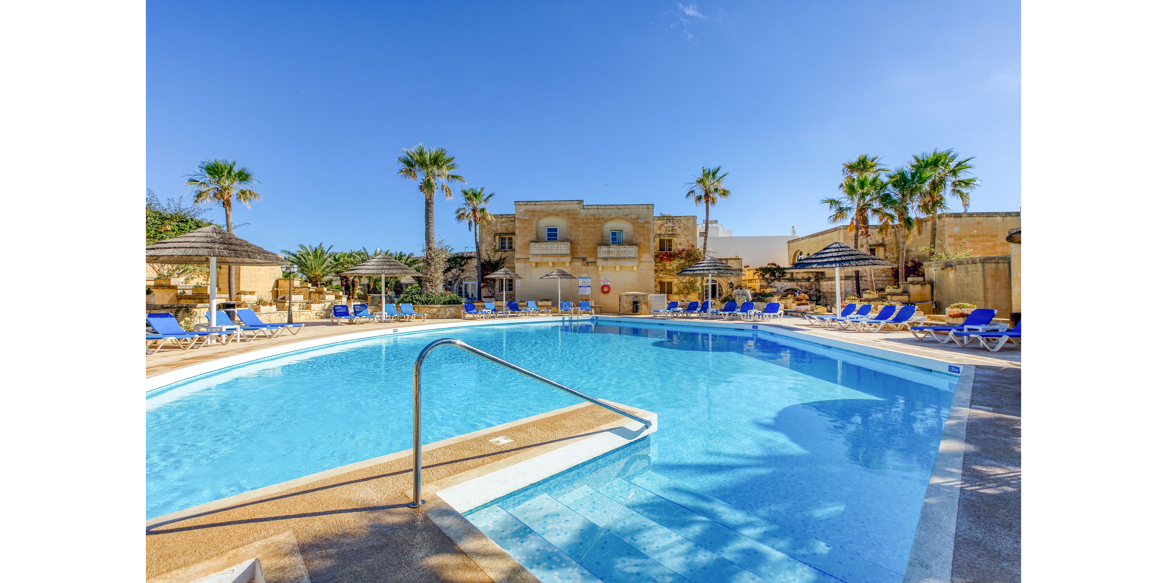 A large outdoor swimming pool sits at the center of a resort courtyard under a clear blue sky. Blue sun loungers and umbrellas line the poolside, suggesting a relaxing vacation setting. Palm trees and stone buildings surround the area, adding a warm, Mediterranean feel. The calm water and bright sunlight create an inviting atmosphere for leisure and sunbathing.