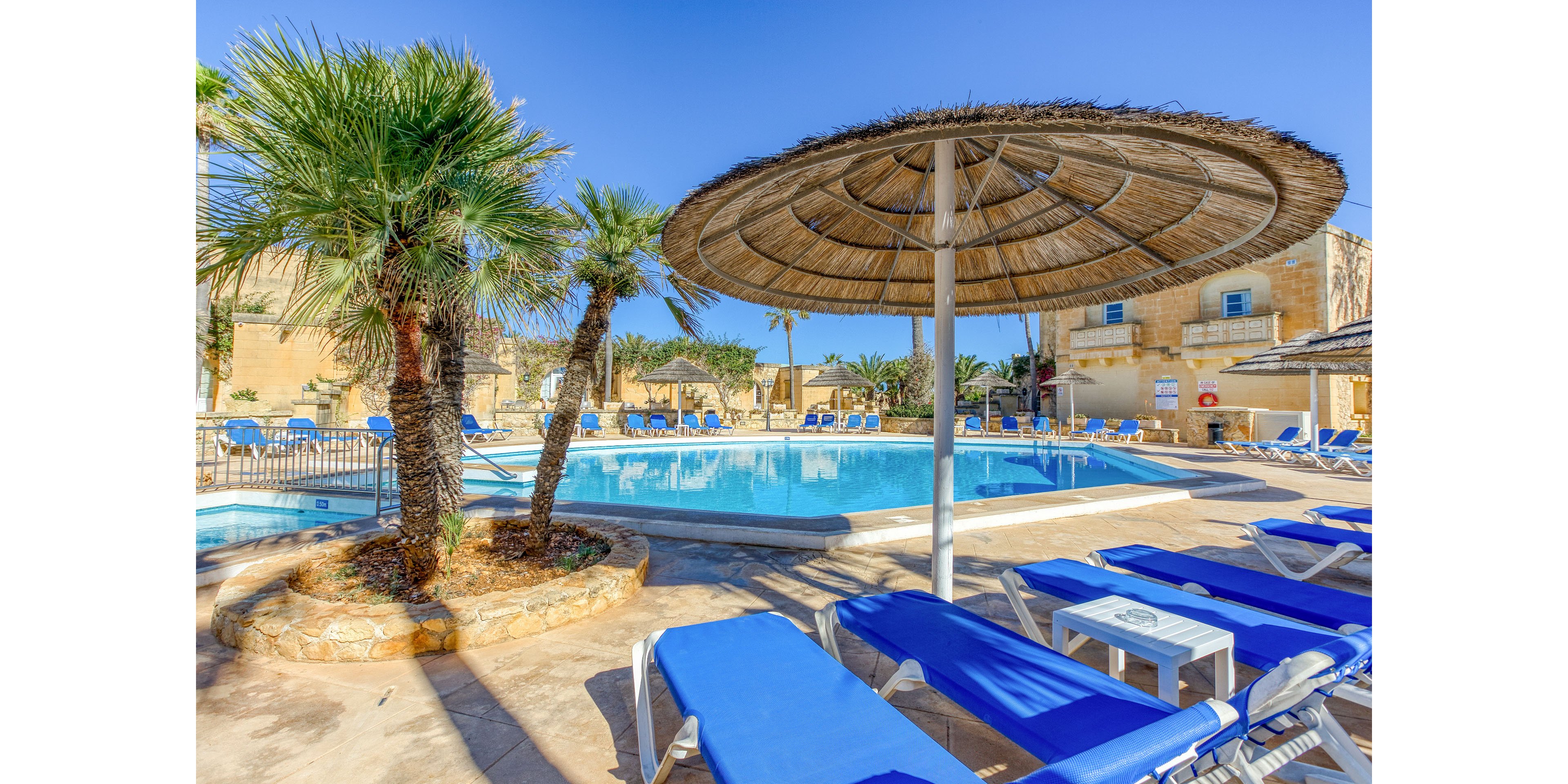 Outdoor resort swimming pool surrounded by blue sun loungers and straw parasols under a clear blue sky. Palm trees and stone walls create a Mediterranean vacation atmosphere. The bright sunlight highlights the calm water and relaxed seating area. No people are present, emphasizing a peaceful and inviting holiday setting.