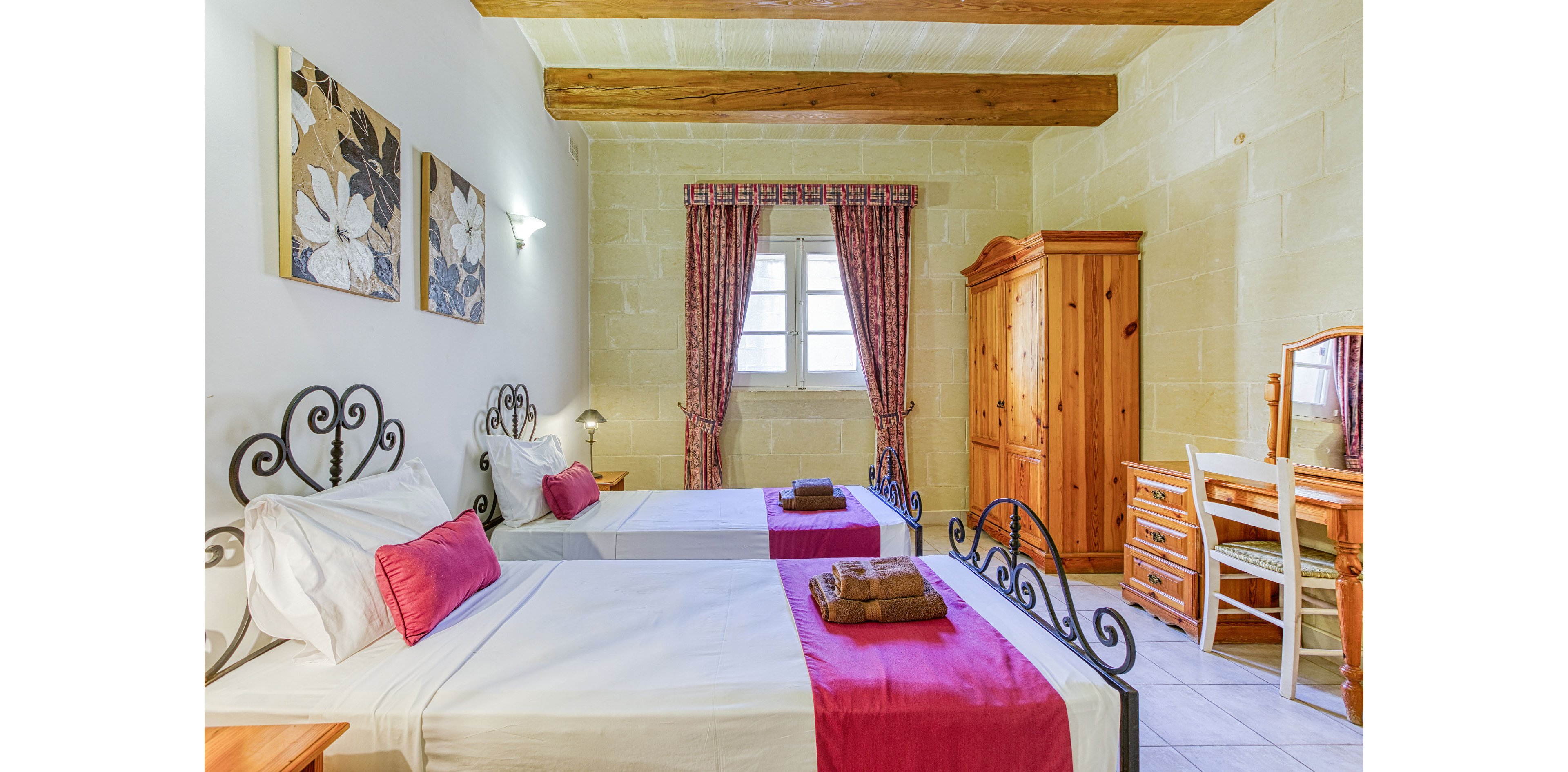 Bright twin bedroom featuring two single beds with white linens and magenta runners and cushions. The room has stone walls, exposed wooden ceiling beams, and a tiled floor. A wooden wardrobe and matching desk with mirror sit opposite the beds, while folded towels rest neatly on each bed. Floral wall art and patterned curtains frame the window, adding a warm, inviting touch to the space.