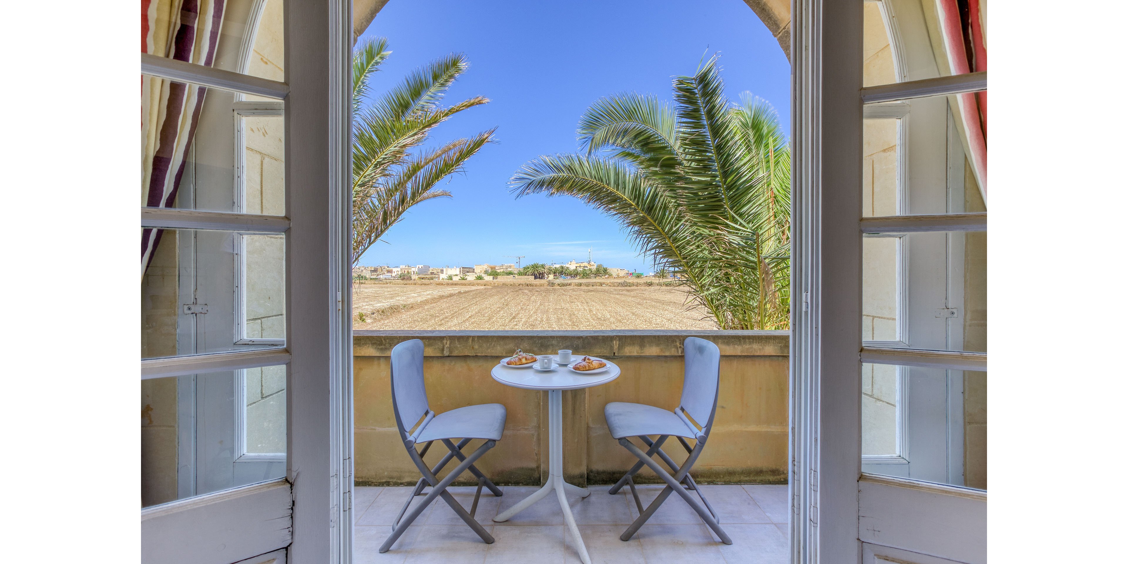 A small balcony table is set for breakfast with two chairs facing an open countryside view. The scene is framed by open French doors and palm trees, creating a bright and airy atmosphere. Clear blue sky stretches over plowed fields and distant buildings, suggesting a peaceful rural setting. The overall mood is relaxed and inviting, ideal for a quiet morning meal outdoors.