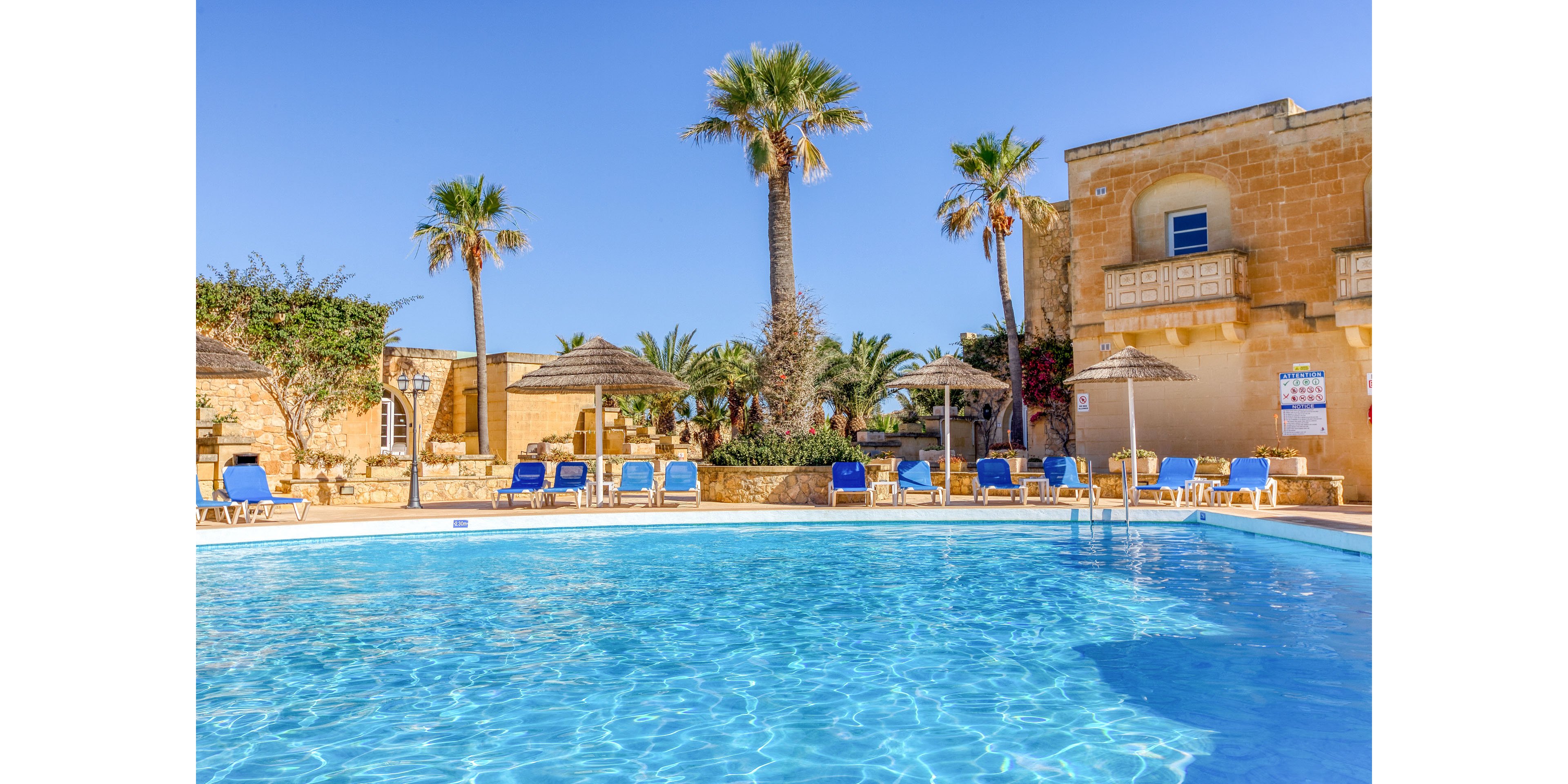 A clear blue swimming pool sits in front of a warm-toned resort building under a bright, cloudless sky. Palm trees and lush greenery surround the pool, creating a tropical holiday atmosphere. Blue sun loungers and straw parasols line the poolside, suggesting a relaxing vacation setting. The scene is brightly lit, emphasizing the inviting water and tranquil outdoor space.