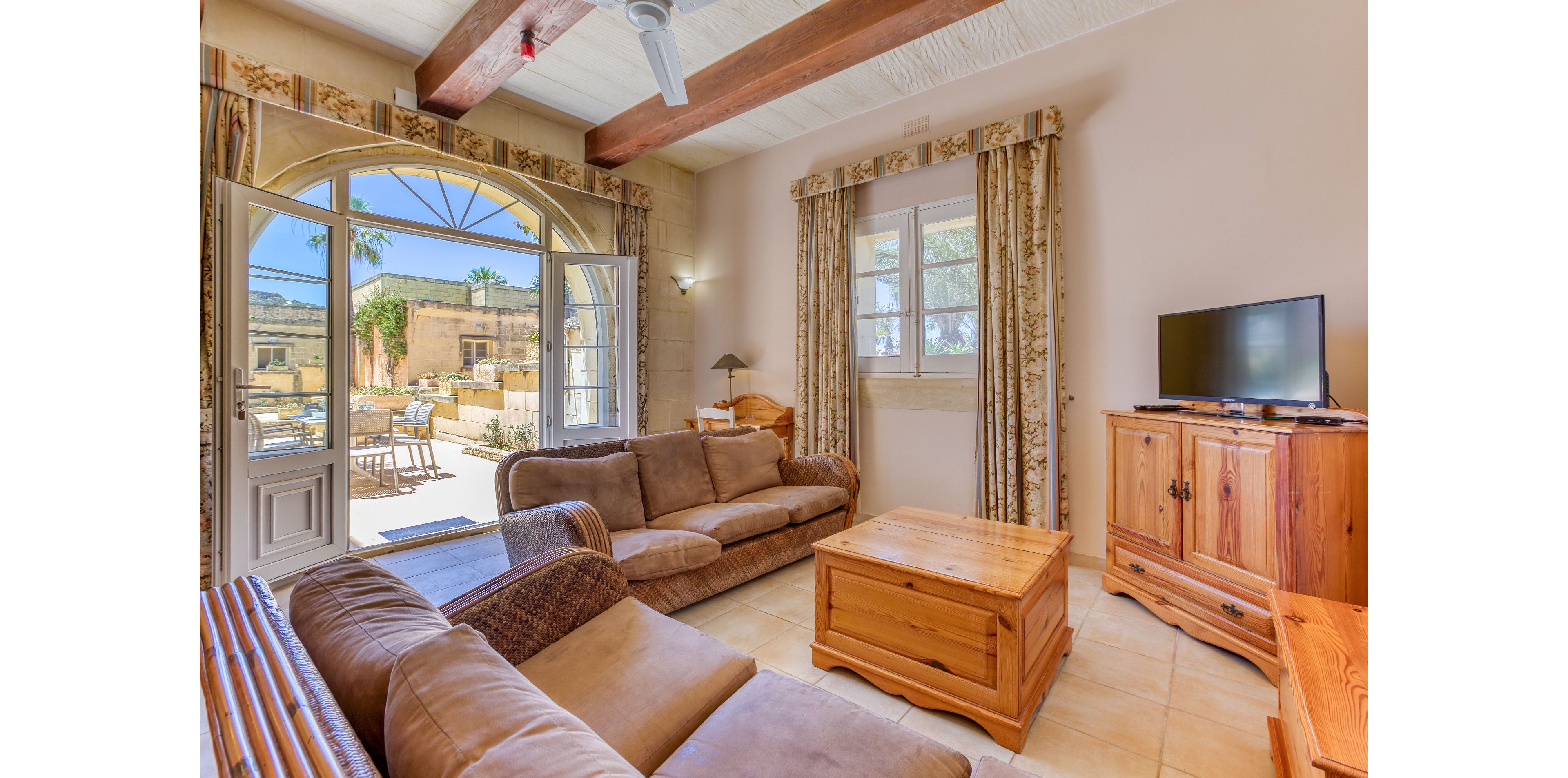 Cozy living room featuring brown upholstered sofas and wooden furniture in a rustic style. Large arched French doors open to a sunny outdoor patio with seating and palm trees visible outside. Exposed wooden ceiling beams and patterned curtains add warmth and character to the space. A flat-screen television sits on a wooden cabinet, completing the comfortable holiday home atmosphere.
